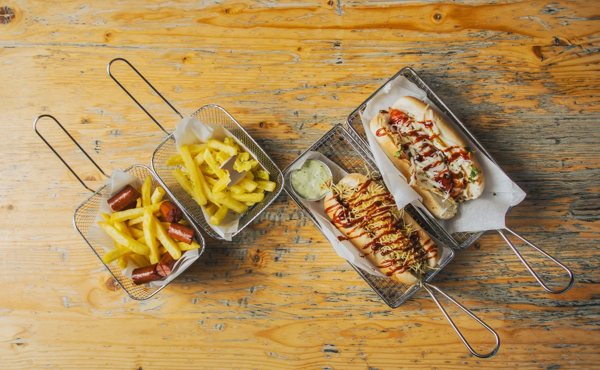 Porções de batatas e salsichas na esquerda, hot dogs com molhos e acompanhamentos na direita, tudo servido em cestas de metal sobre uma mesa de madeira.