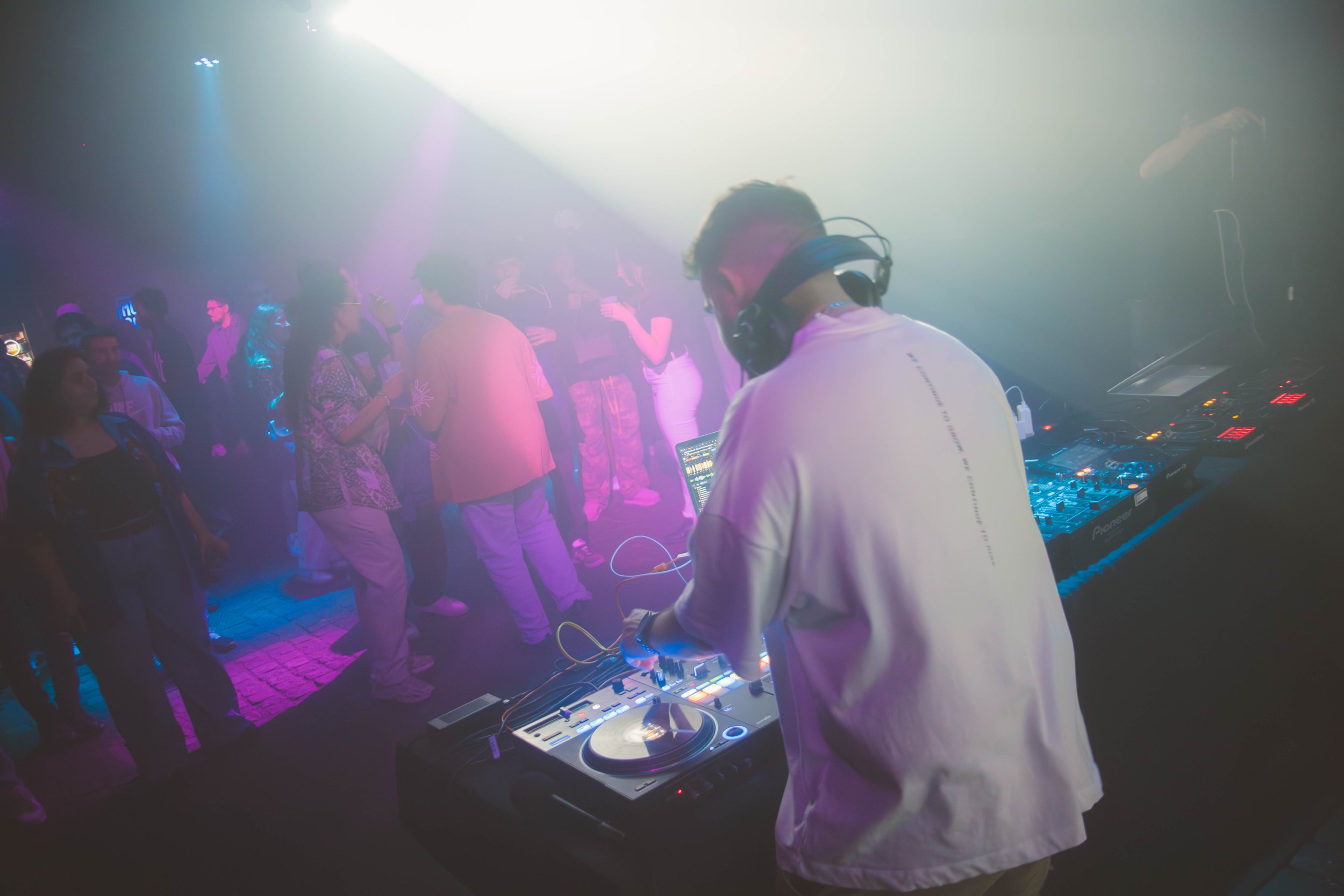 DJ em um clube noturno tocando músicas com pessoas dançando ao fundo, luzes coloridas e atmosfera de festa.