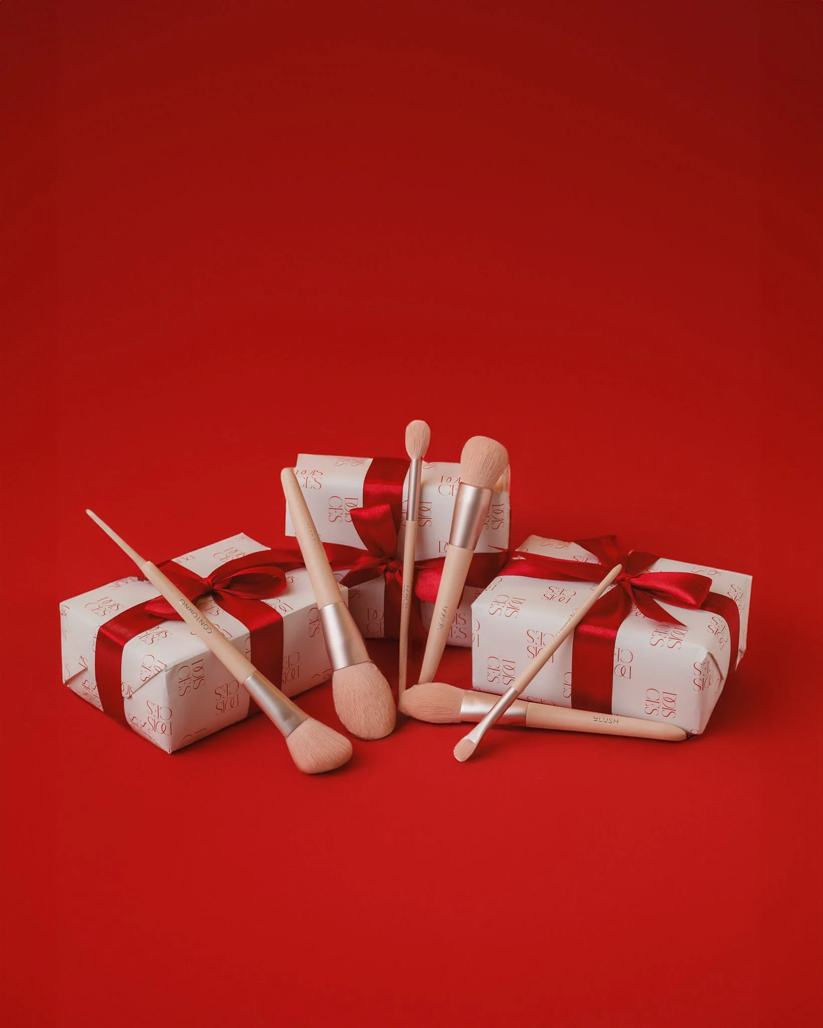 Conjunto de cinco presentes embalados com papel branco, fita vermelha, decorados com laço vermelho, ao lado de sete pincéis de maquiagem com cabos brancos e cerdas rosa claro, sobre fundo vermelho.