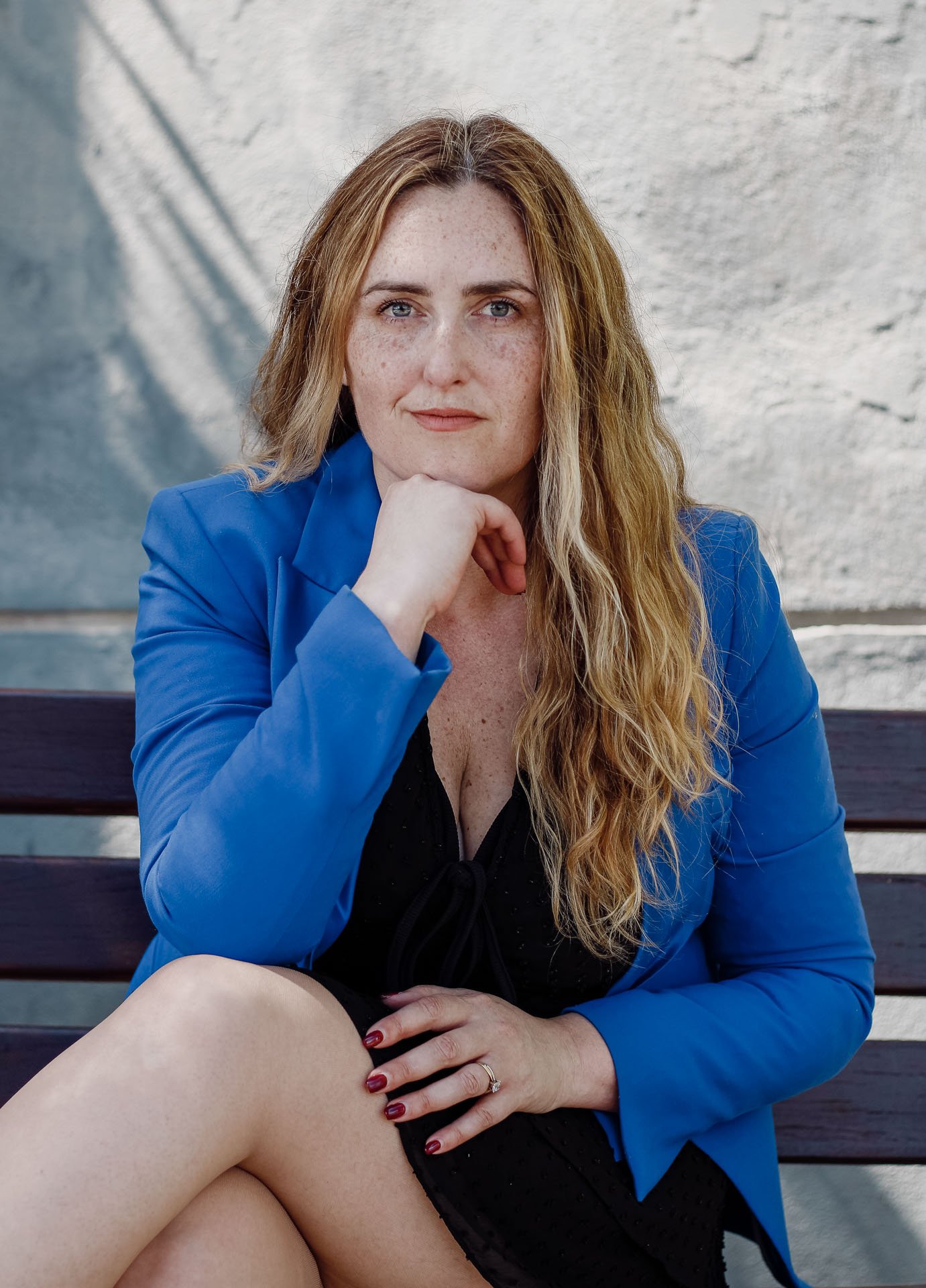 Mulher de cabelos ruivos, olhos azuis, usando blazer azul escuro, sentado em um banco de madeira, com cabelo ondulado, usando vestido preto e unha pintada de vinho, olhando para a câmera com expressão séria.