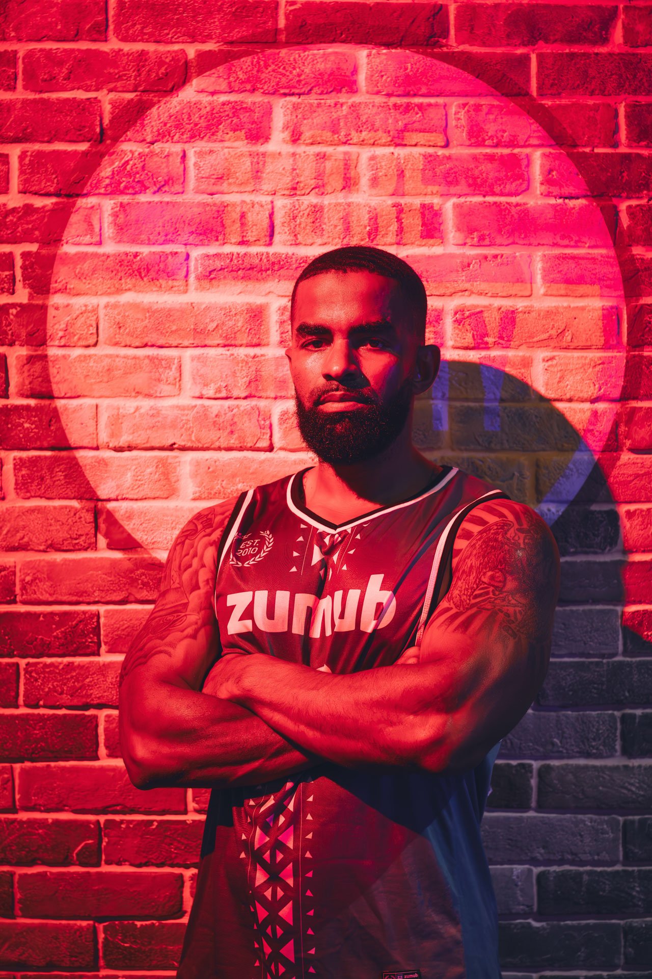 Homem com barba, braços cruzados, vestindo camisa de time Zumba, em frente a parede de tijolos vermelhos com iluminação vermelha