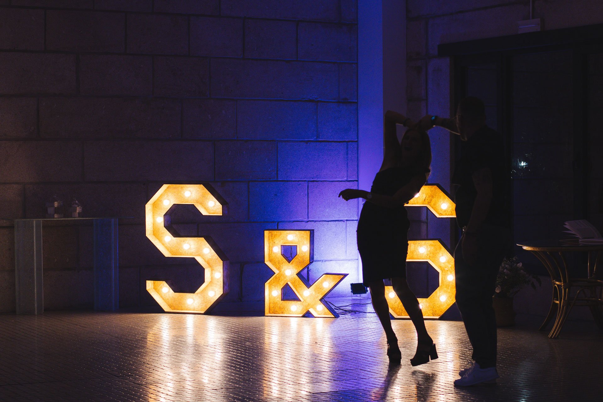 Silhuetas de um casal dançando ao lado de letras iluminadas S & L em um fundo escuro de parede de blocos de cimento.