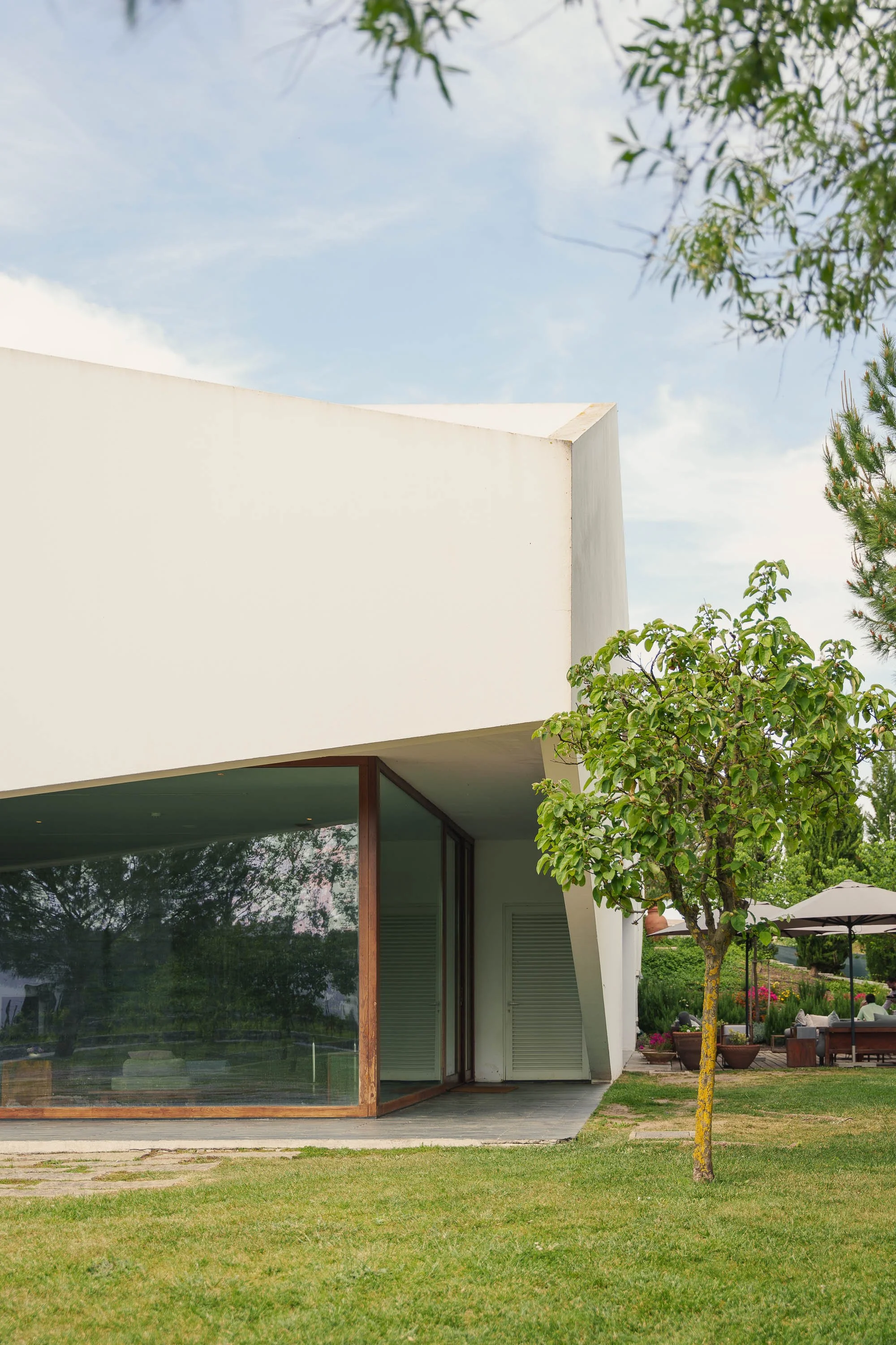 Casa moderna branca com grandes janelas de vidro, cercada por árvores e gramado verde, sob céu parcialmente nublado.