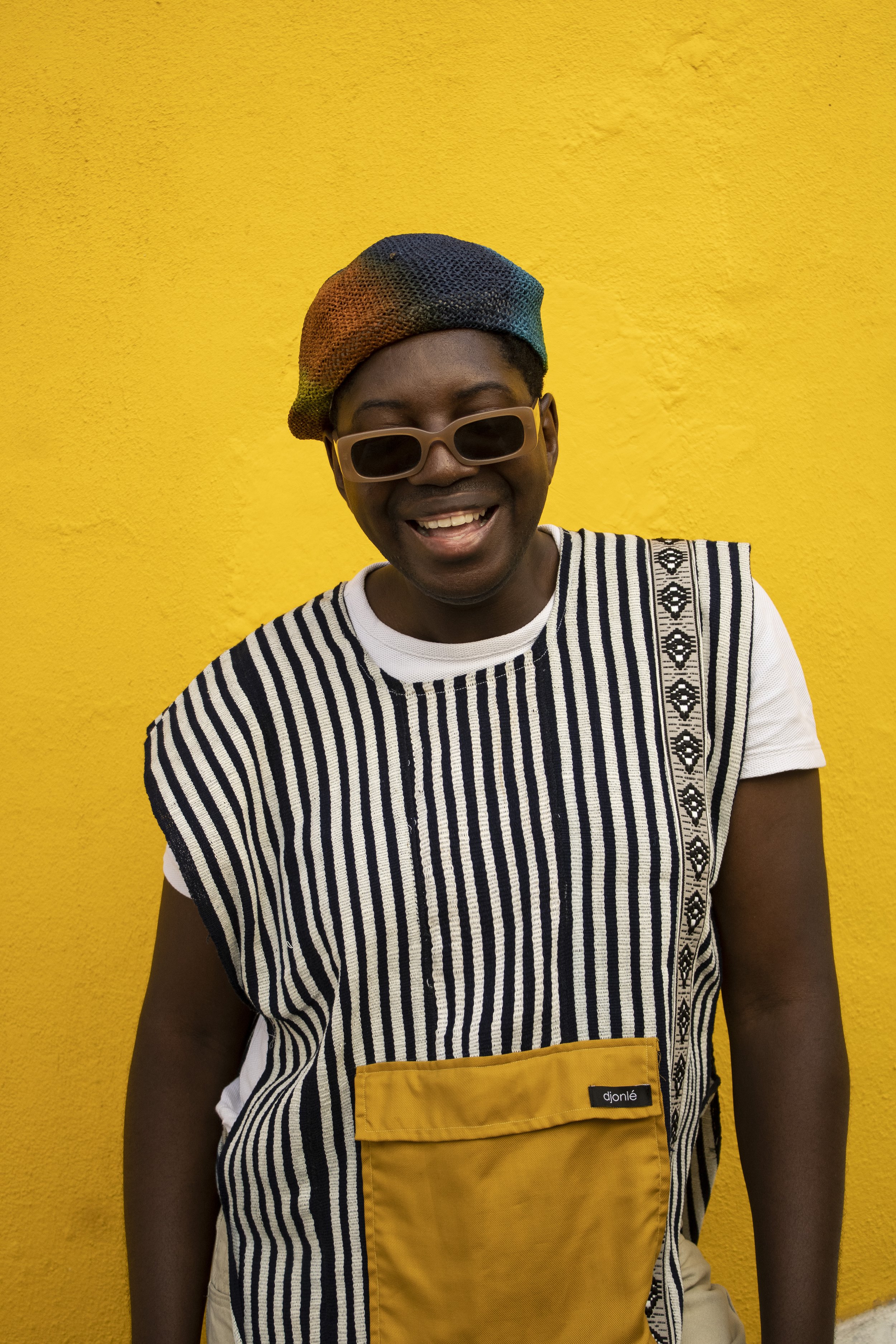 Pessoa sorridente com óculos escuros, chapéu colorido e blusa de riscas, posando contra parede amarela.