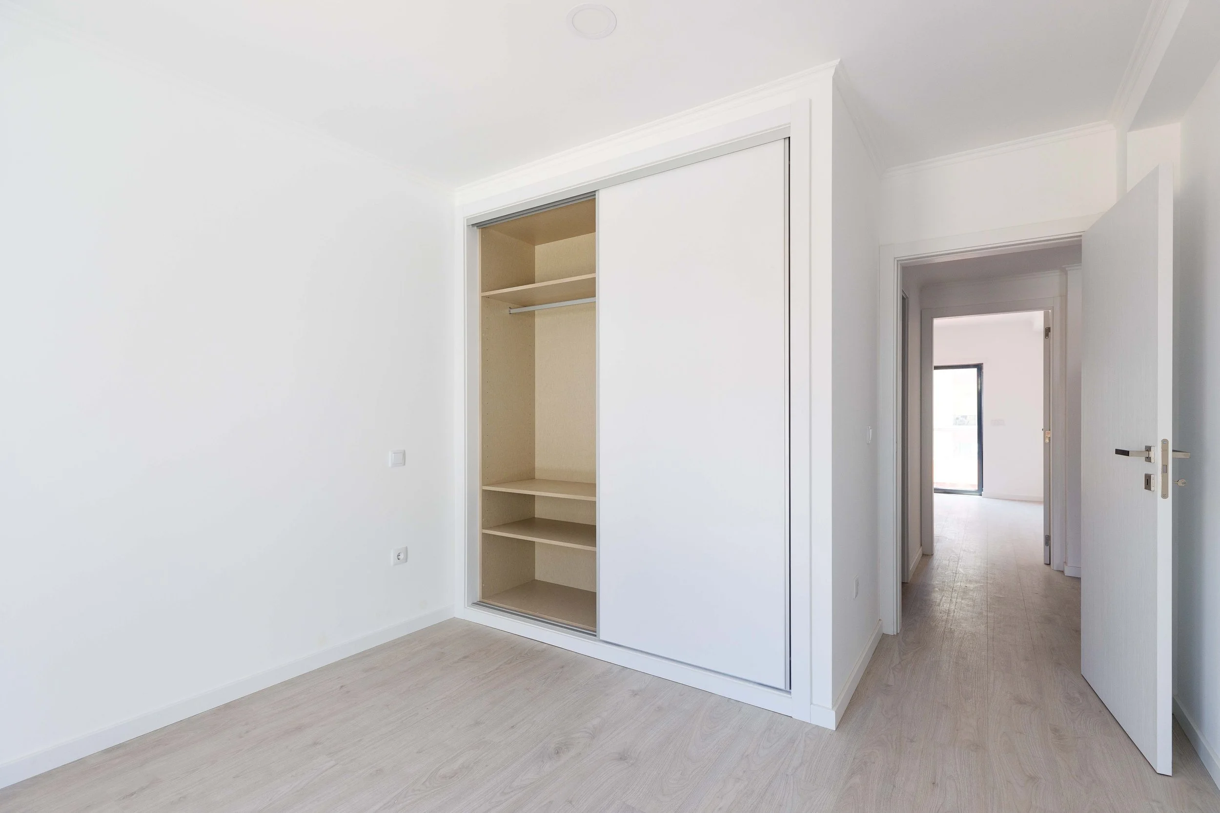 Quarto vazio com armário embutido de portas brancas e piso de madeira clara, portas abertas levando a outros cômodos.
