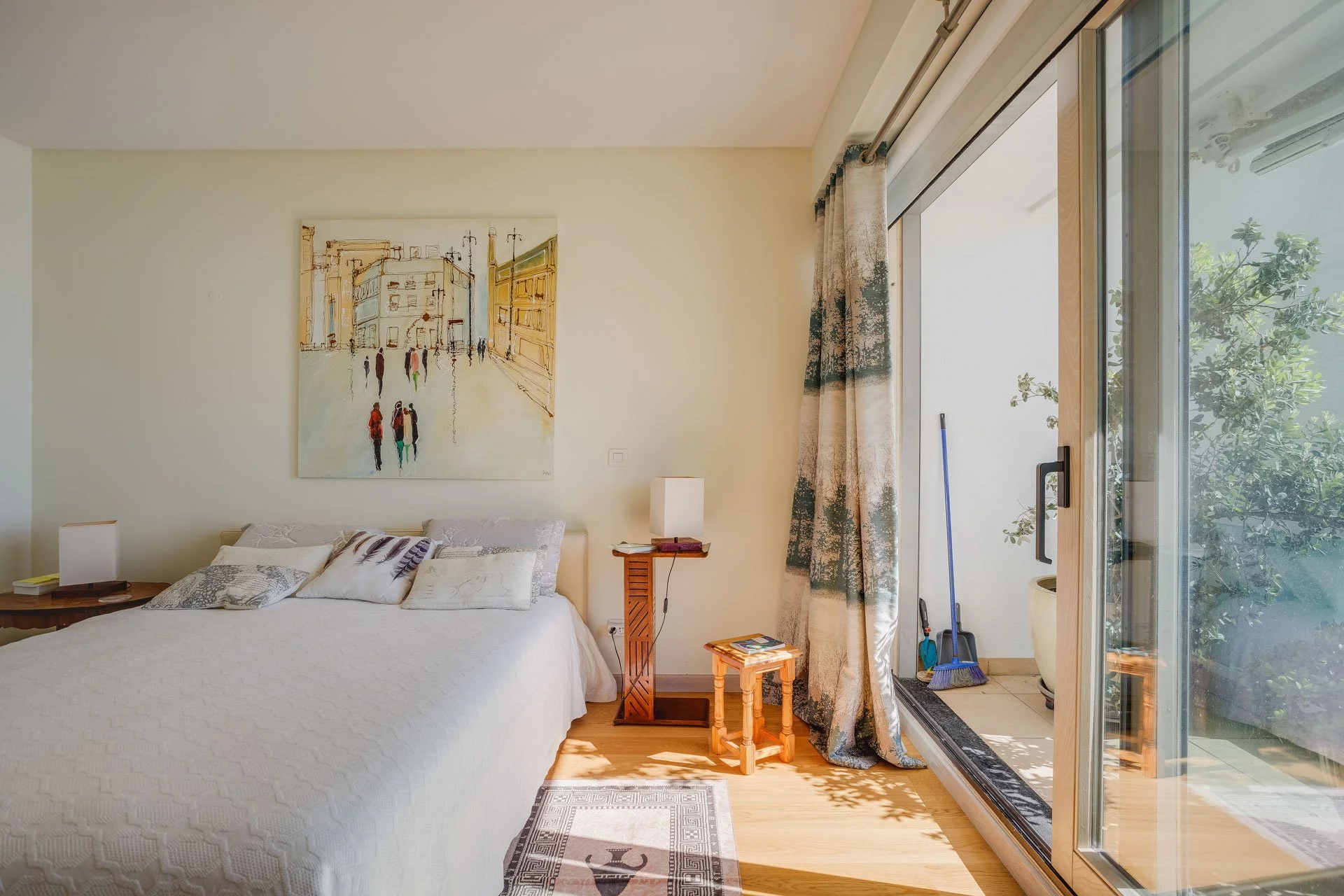 Quarto com cama de cama branca, arte na parede de uma cidade, iluminação natural, vidro de sacada com vista para plantas e ferramentas de limpeza.