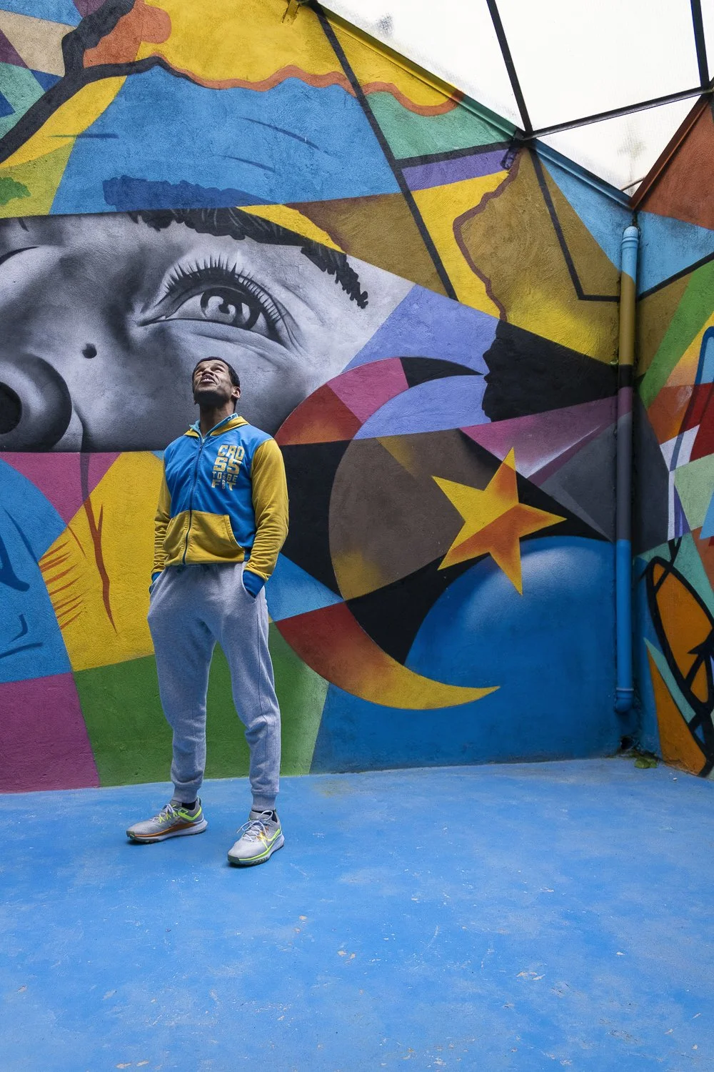 Homem posando à frente de uma parede de arte urbana colorida com um retrato de rosto de mulher, estrelas, luas, formas geométricas e cores vibrantes.