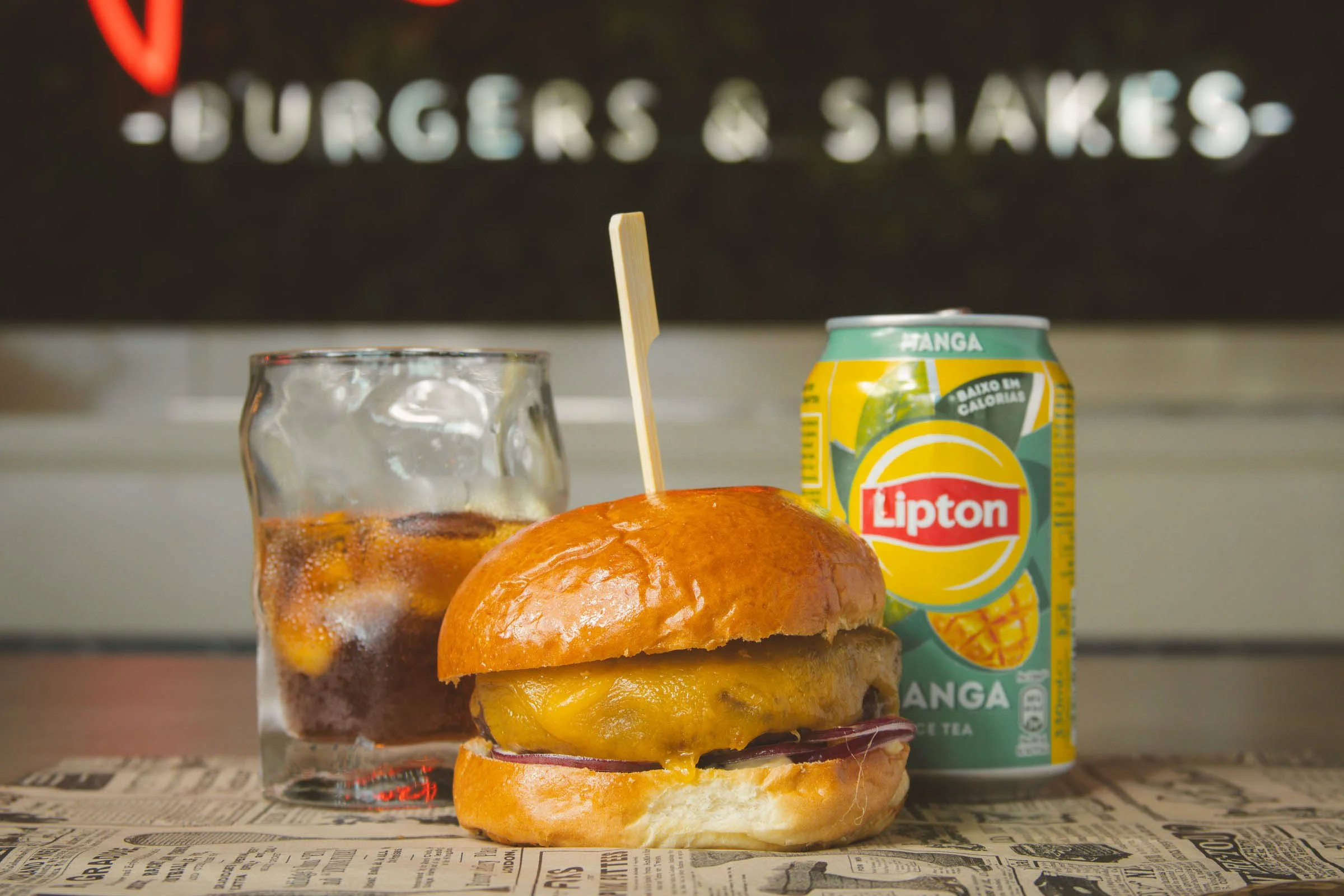 Sanduíche com pão, queijo derretido, carne e cebola, acompanhado de uma lata de refrigerante Lipton de sabor manga e um copo com bebida e gelo, sobre uma mesa com papel de jornal, ao fundo um letreiro com as palavras 'Burgers & Shakes'.