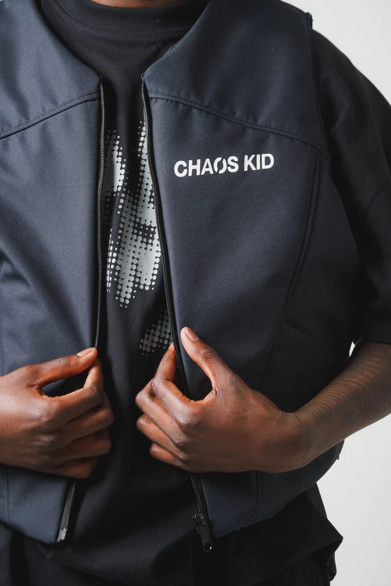 Pessoa usando jaqueta preta com a inscrição 'CHAOS KID' e camiseta preta com um desenho de pontos brancos, ajustando a jaqueta