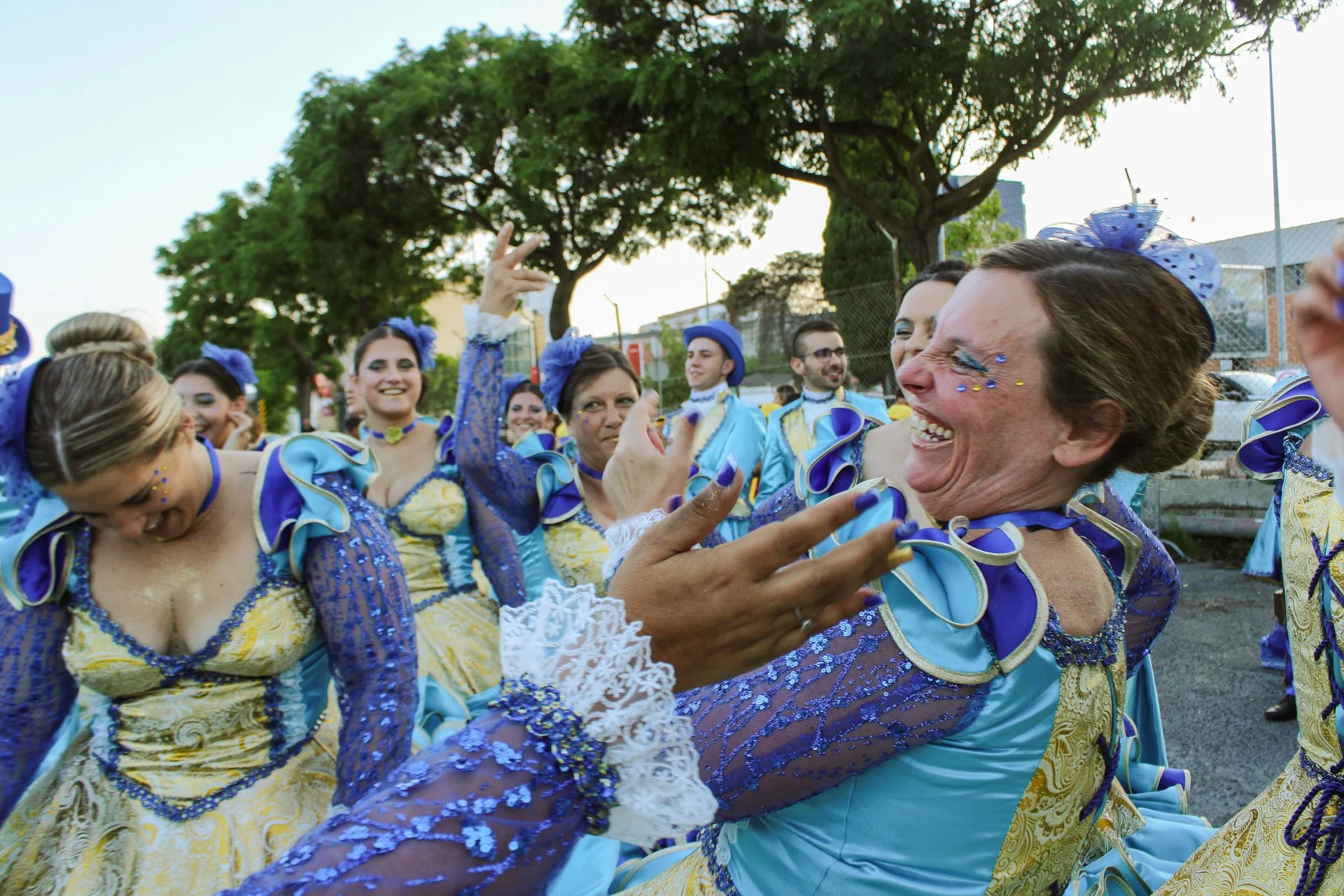 Mulheres em fantasias azuis e amarelas comemorando e dançando, uma sorrindo e outra abaixada, em uma celebração ao ar livre com árvores ao fundo.