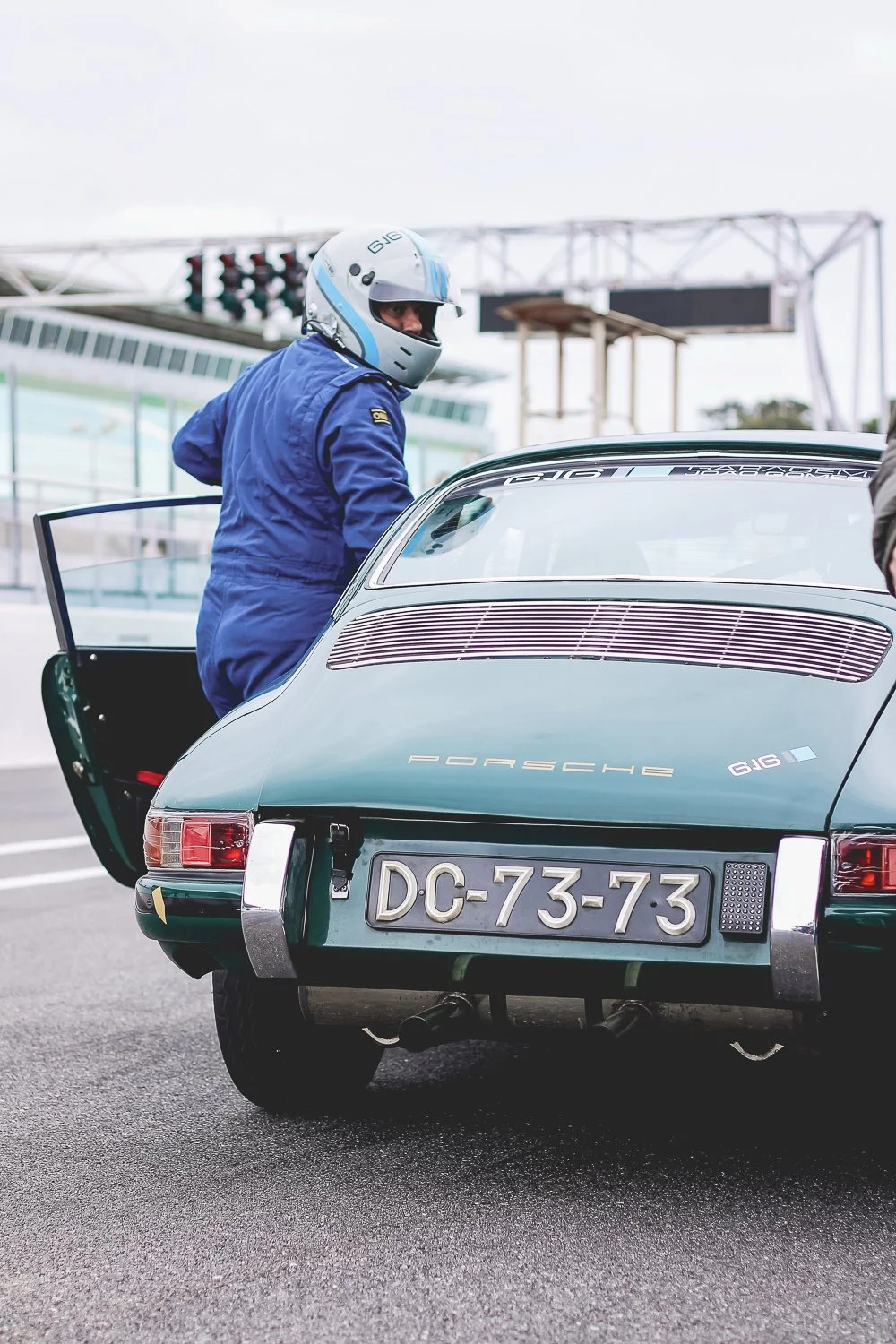 Piloto de capacete azul e jaqueta azul ao lado de um carro de corrida Porsche 911 com placa DC-73-73 na pista de corrida.
