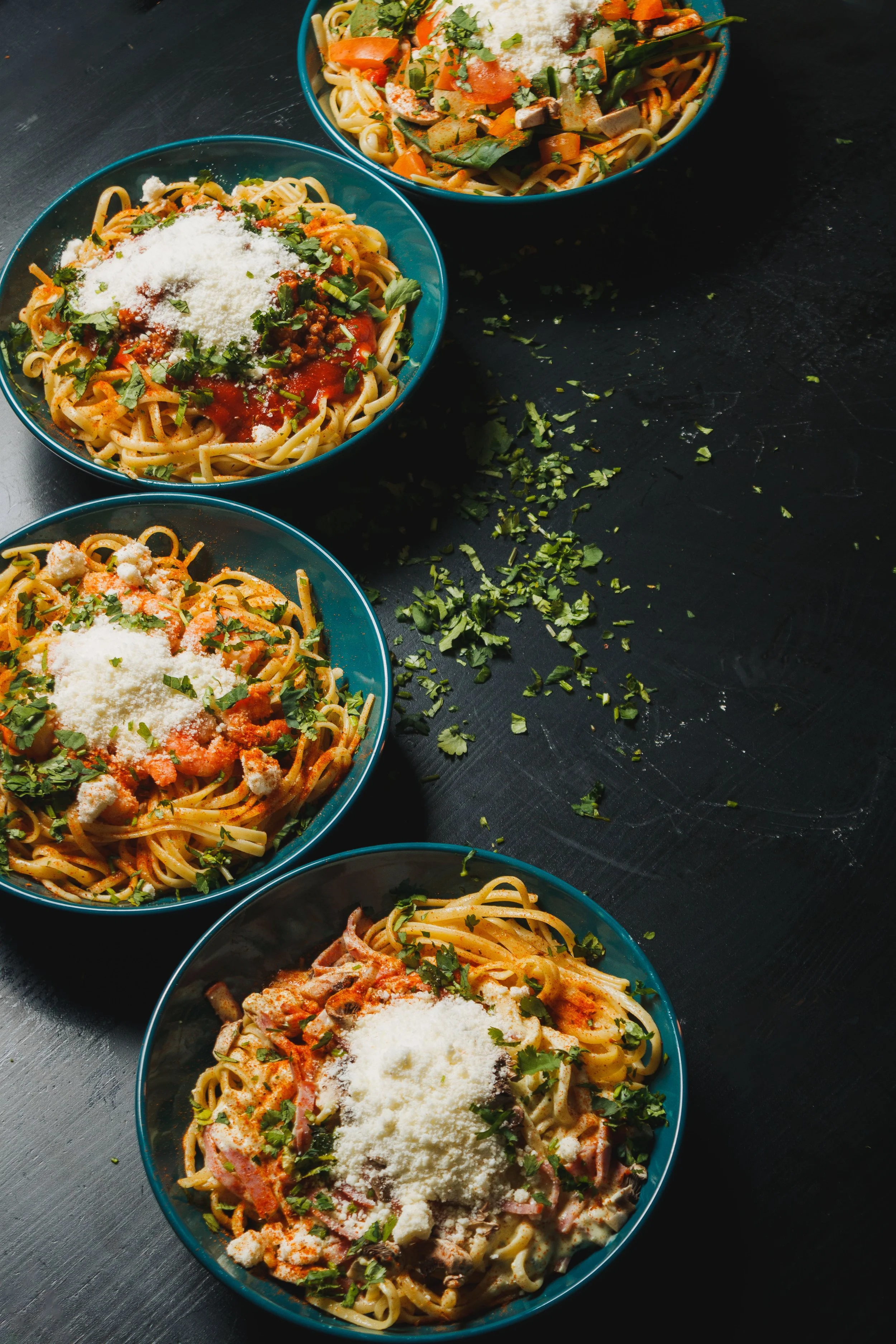 Quatro tigelas de espaguete com molho, queijo ralado e ervas em uma mesa preta, decoradas com tomate, carne, frango ou ingredientes variados.