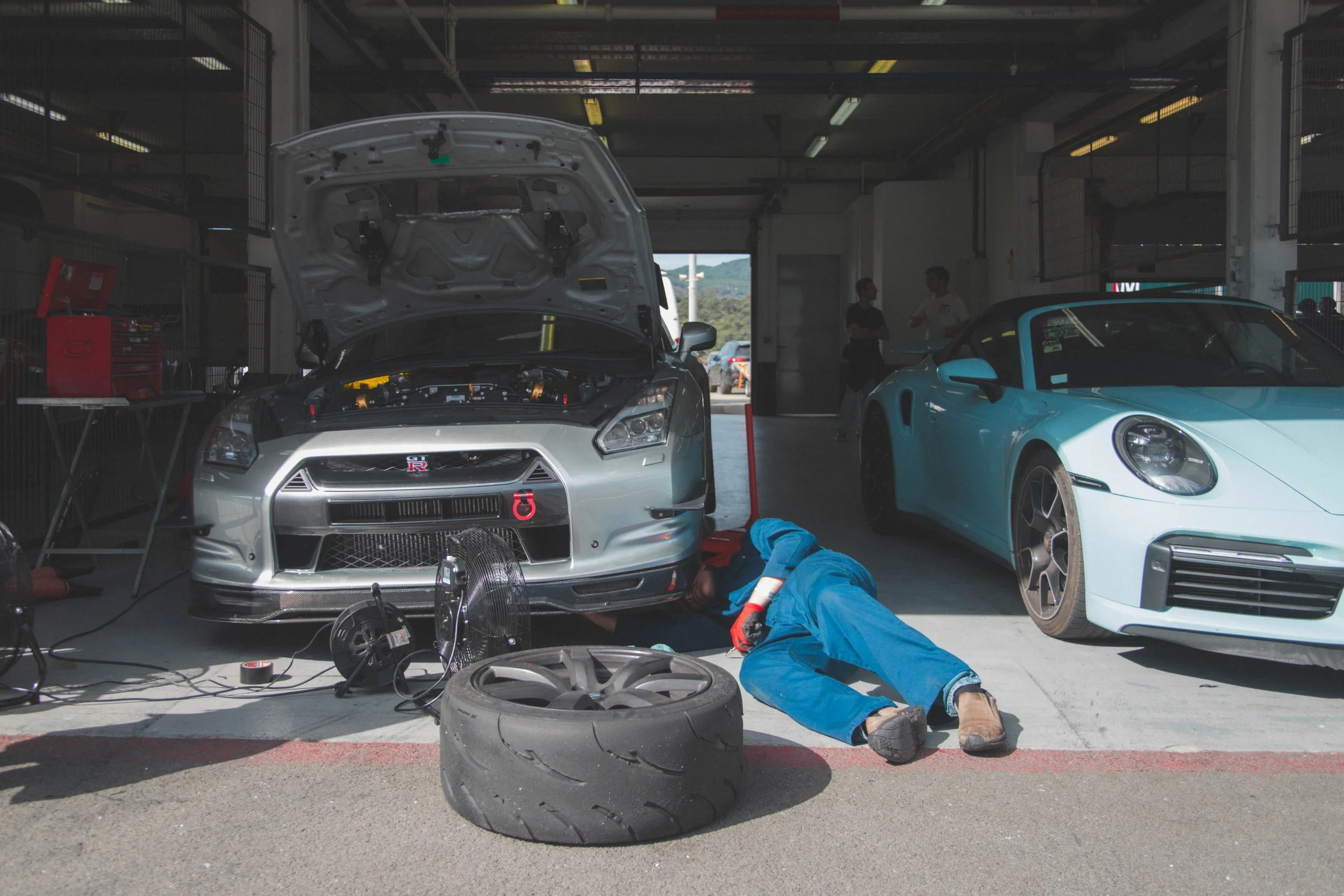 Homem em uma oficina de carros, deitado de bruços verificando o motor de um Nissan GT-R com o capô aberto, próximo a um pneu velho e uma ventoinha, com um carro esportivo azul atrás.