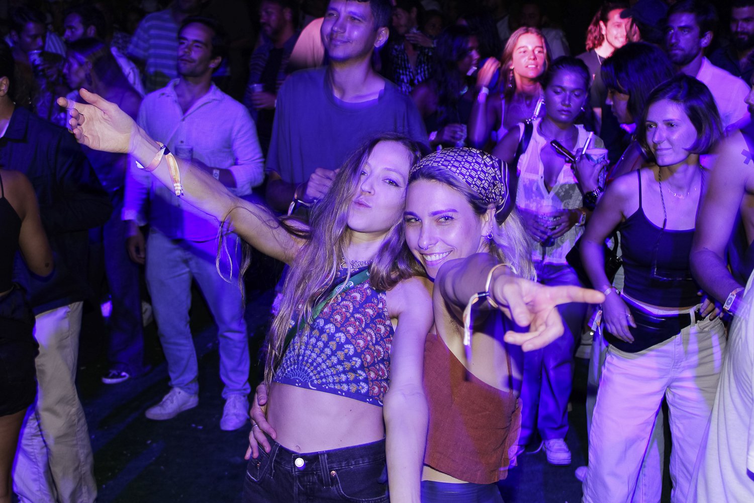 Pessoas dançando e se divertindo em uma festa com luzes coloridas, com destaque para duas mulheres na frente fazendo poses animadas.