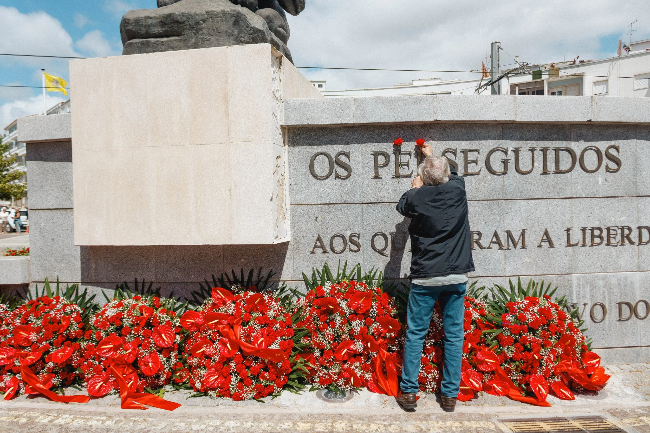 Pessoa colocando flores vermelhas em uma lápide de pedra, com a frase "OS PESSUGIDOS" escrita nela, em um memorial com flores e uma bandeira ao fundo.