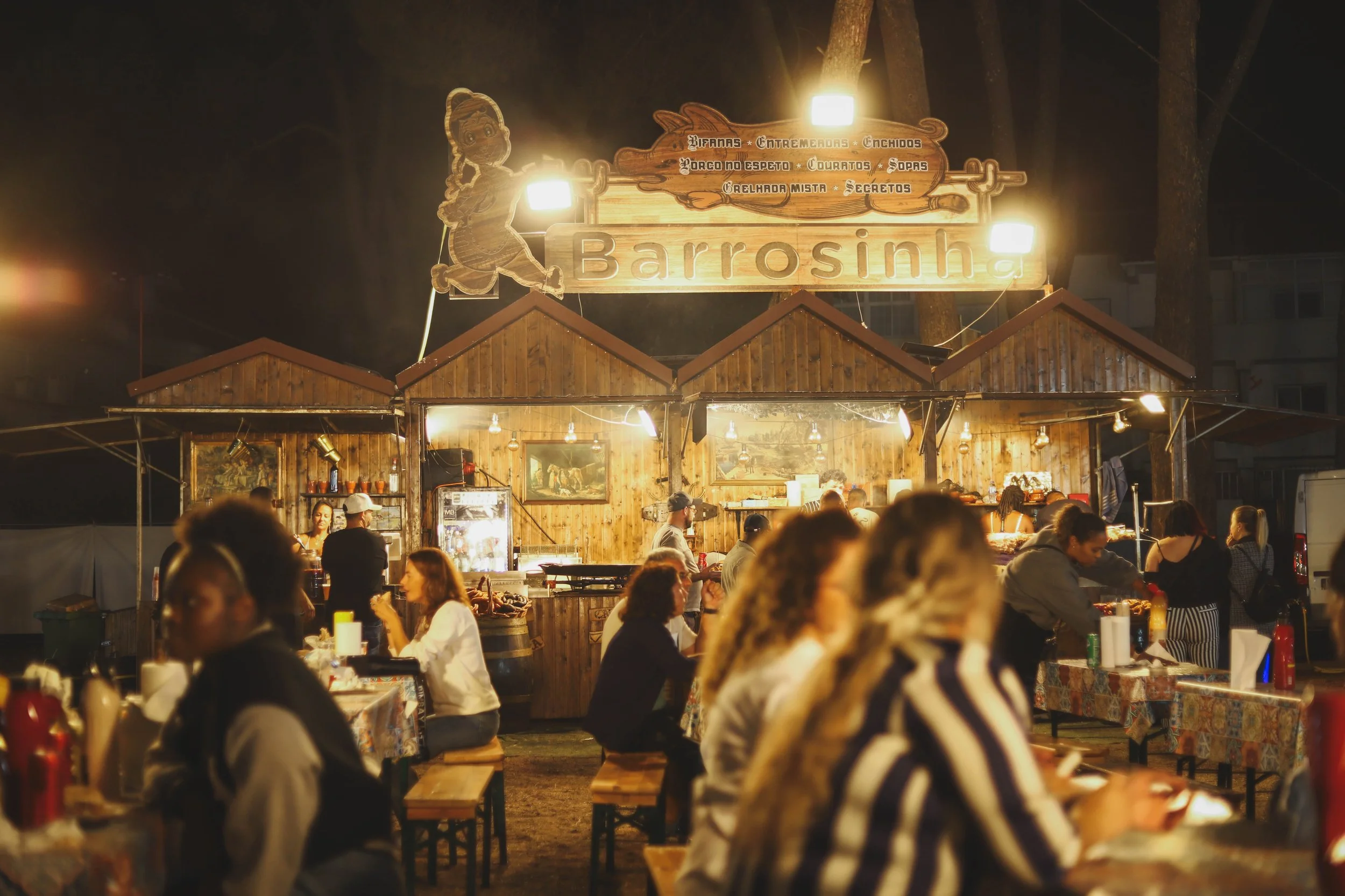 Cena de uma feira noturna ao ar livre com muitas pessoas sentadas em bancos, com barraca de comida de fundo, decorada com madeira e iluminação quente, em uma noite.