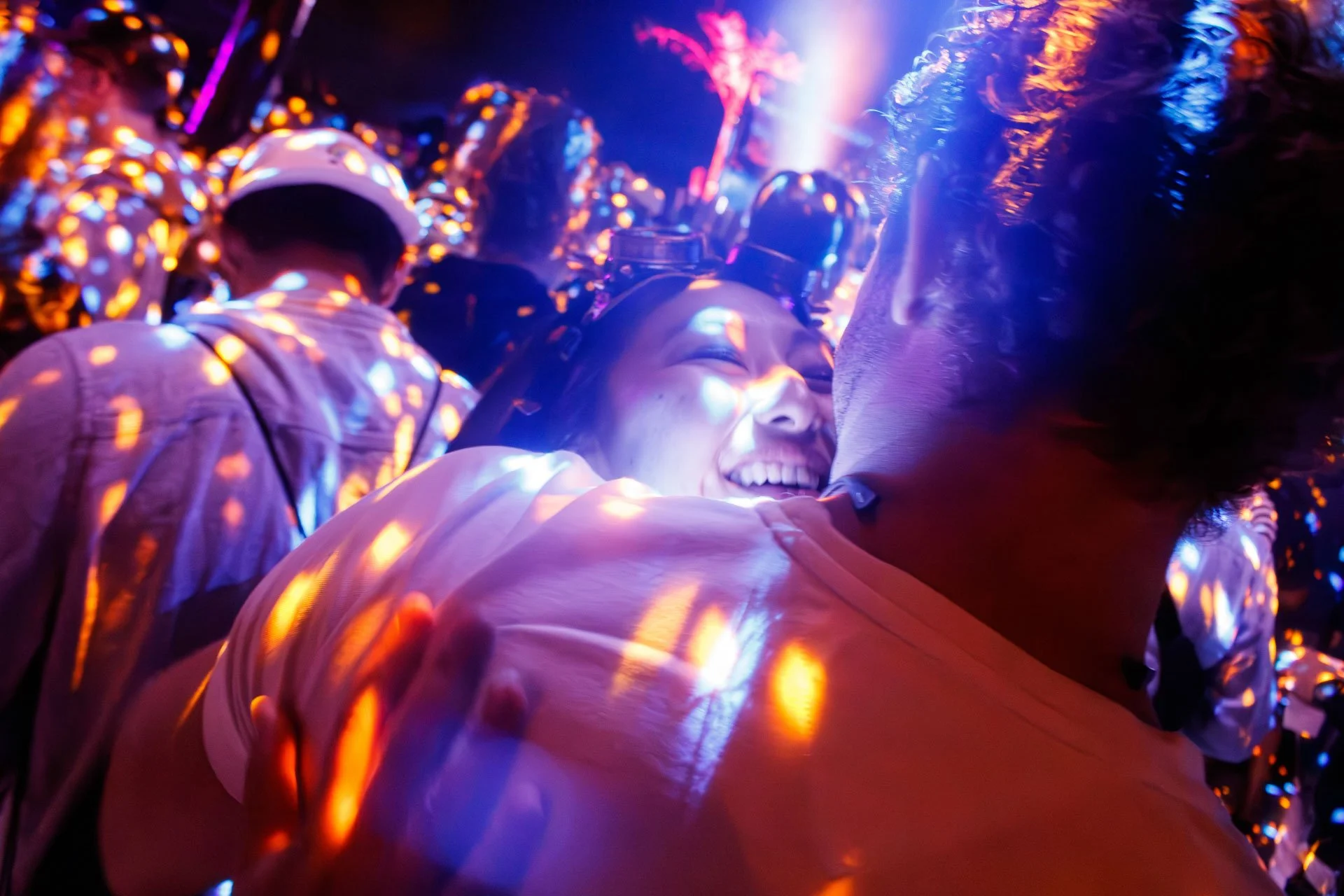 Pessoas em festa com luzes coloridas e reflexos brilhantes, abraçando-se e sorrindo num ambiente animado e festivo.
