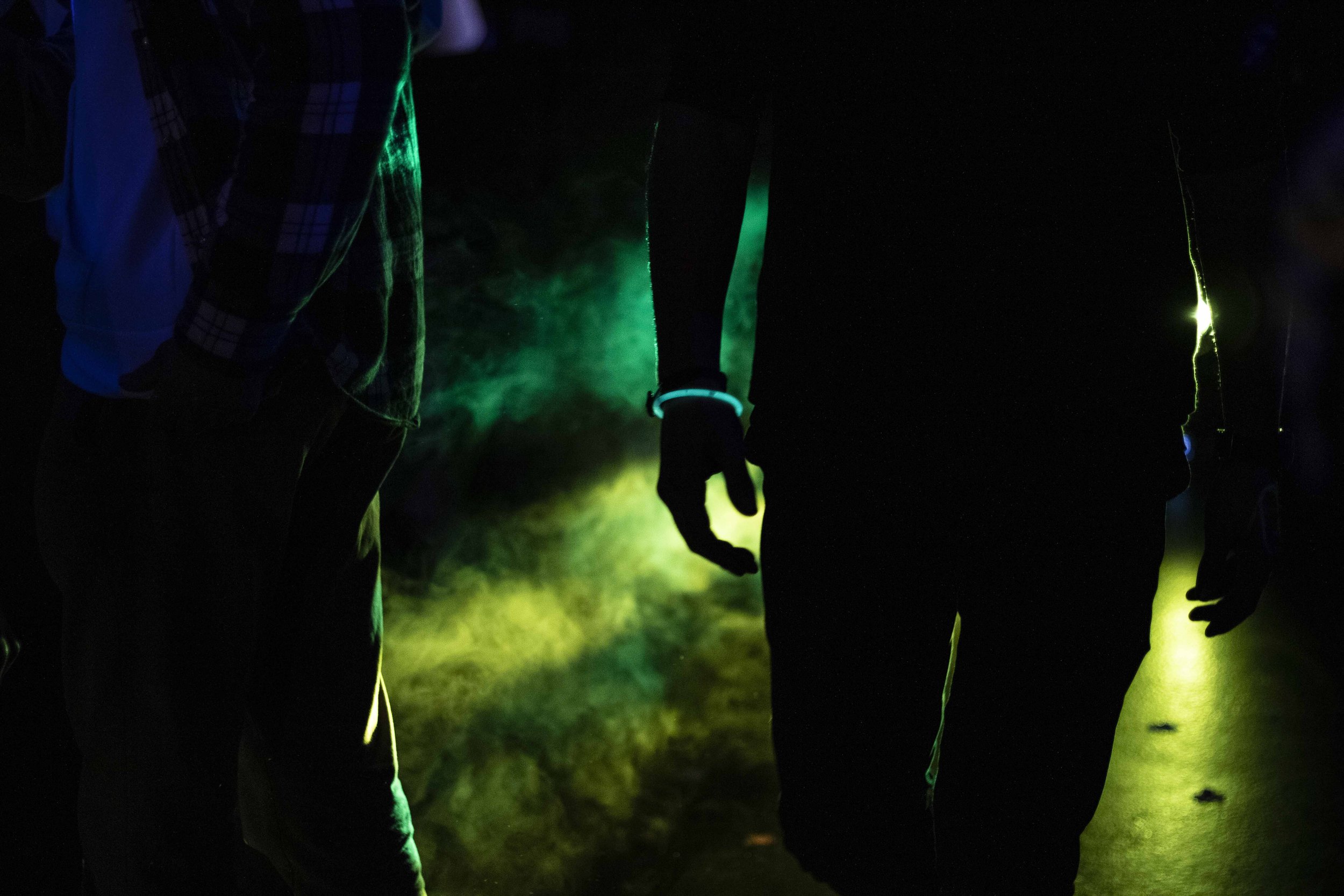 Silhuetas de duas pessoas caminhando em um ambiente escuro com luzes verdes e amarelas ao redor.