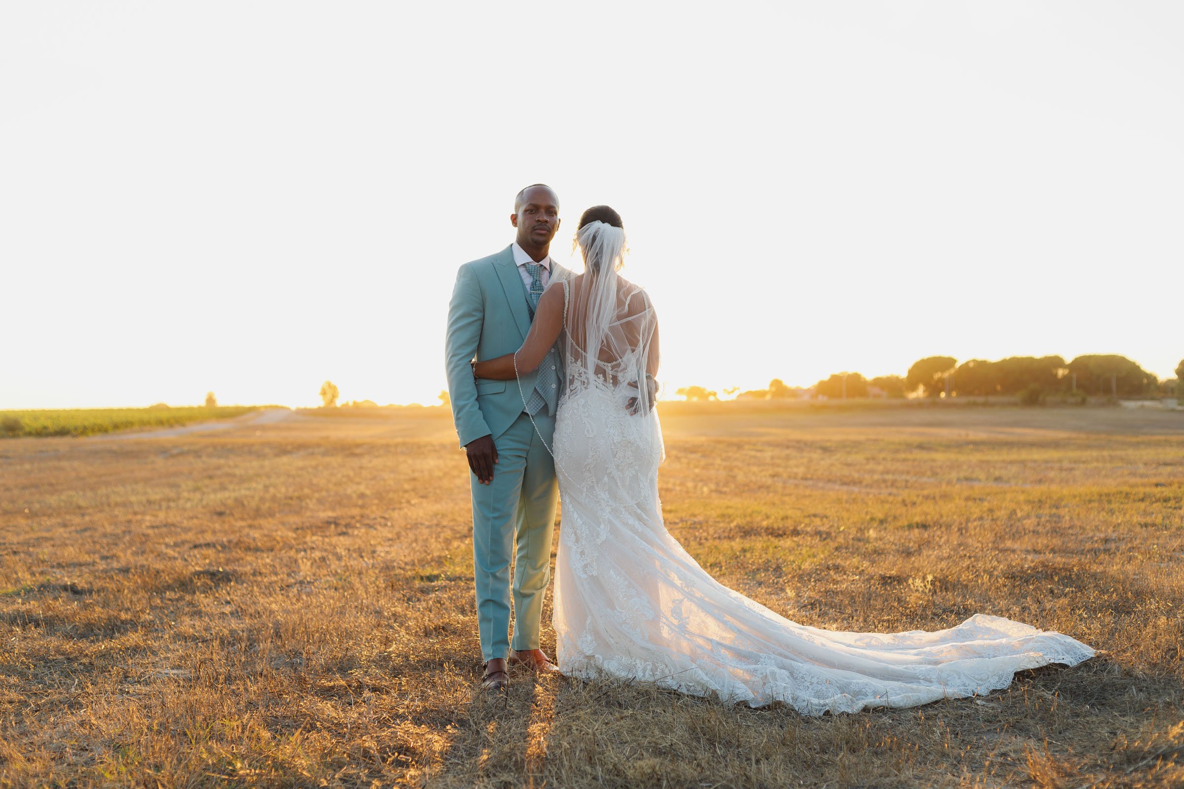 Casal de noivos em campo ao pôr do sol, com ela em vestido de casamento longo e ele em terno azul claro.