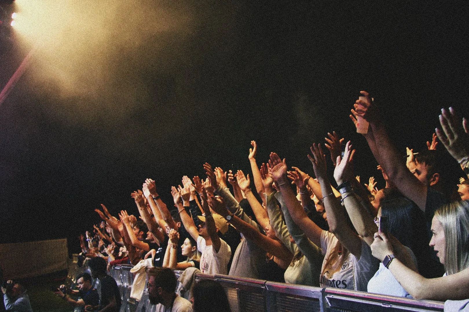 Público em um concerto ou evento com as mãos levantadas na frente de um palco, em um ambiente escuro com luz forte vindo de cima.