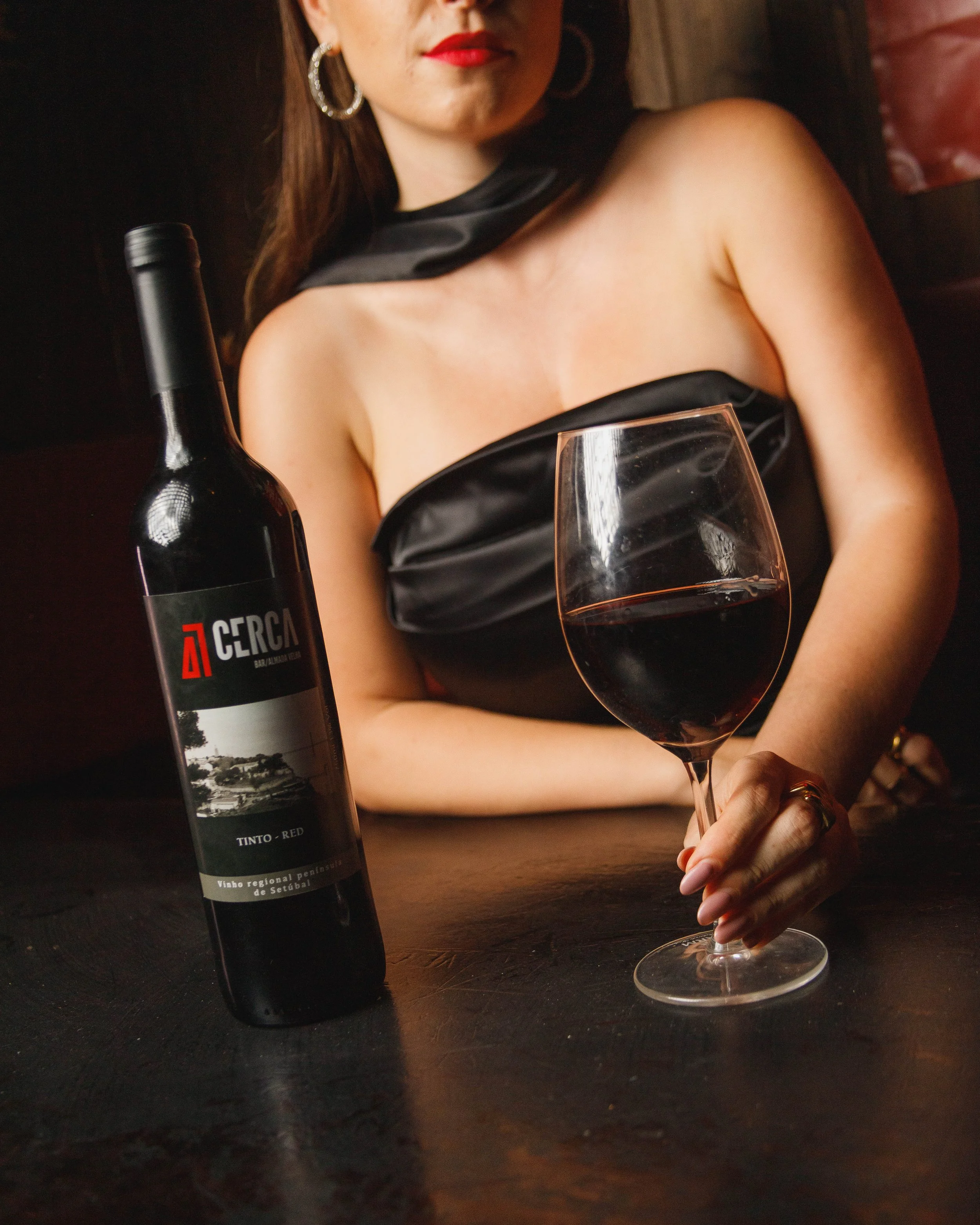 Mulher com lábios vermelhos segurando taça de vinho tinto, ao lado de garrafa de vinho em um ambiente escuro.