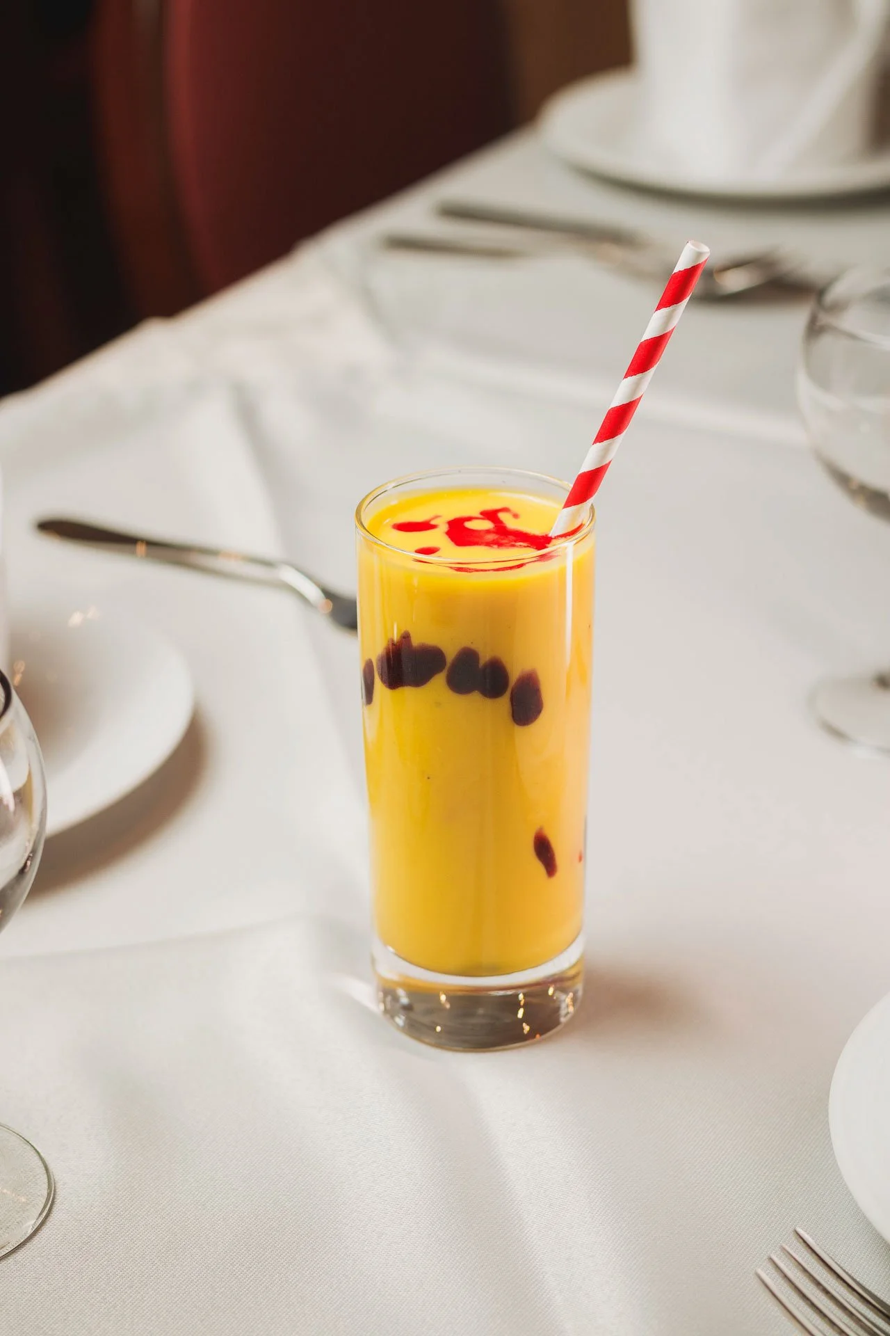 Copo alto com suco de manga, com gelo e gotas de azeite de dendê, com canudo vermelho e branco, colocado sobre uma toalha branca em uma mesa de jantar