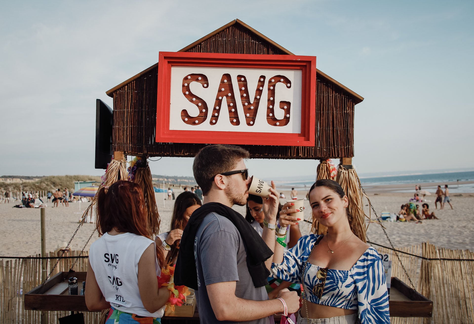 Grupo de pessoas na praia, com uma estrutura de madeira com um painel escrito "SAVG" e algumas pessoas segurando copos.