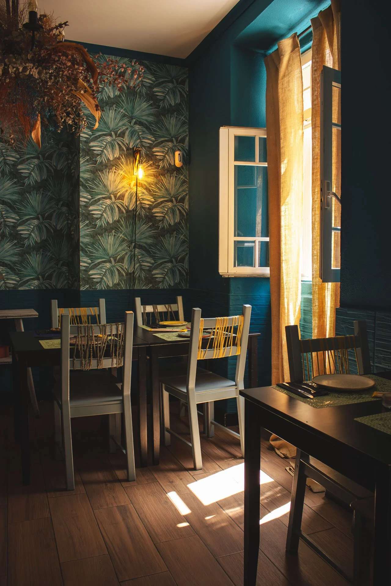 Sala de jantar com janela aberta, cortinas amarelas, parede com papel de parede de folhagens verdes, mesa preta com pratos e talheres e cadeiras brancas e amarelas, iluminação amarelada