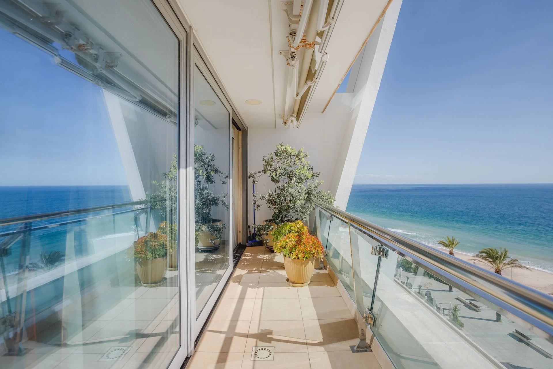 Varanda com vista para o mar, com janelas de vidro, plantas em vasos e céu claro.