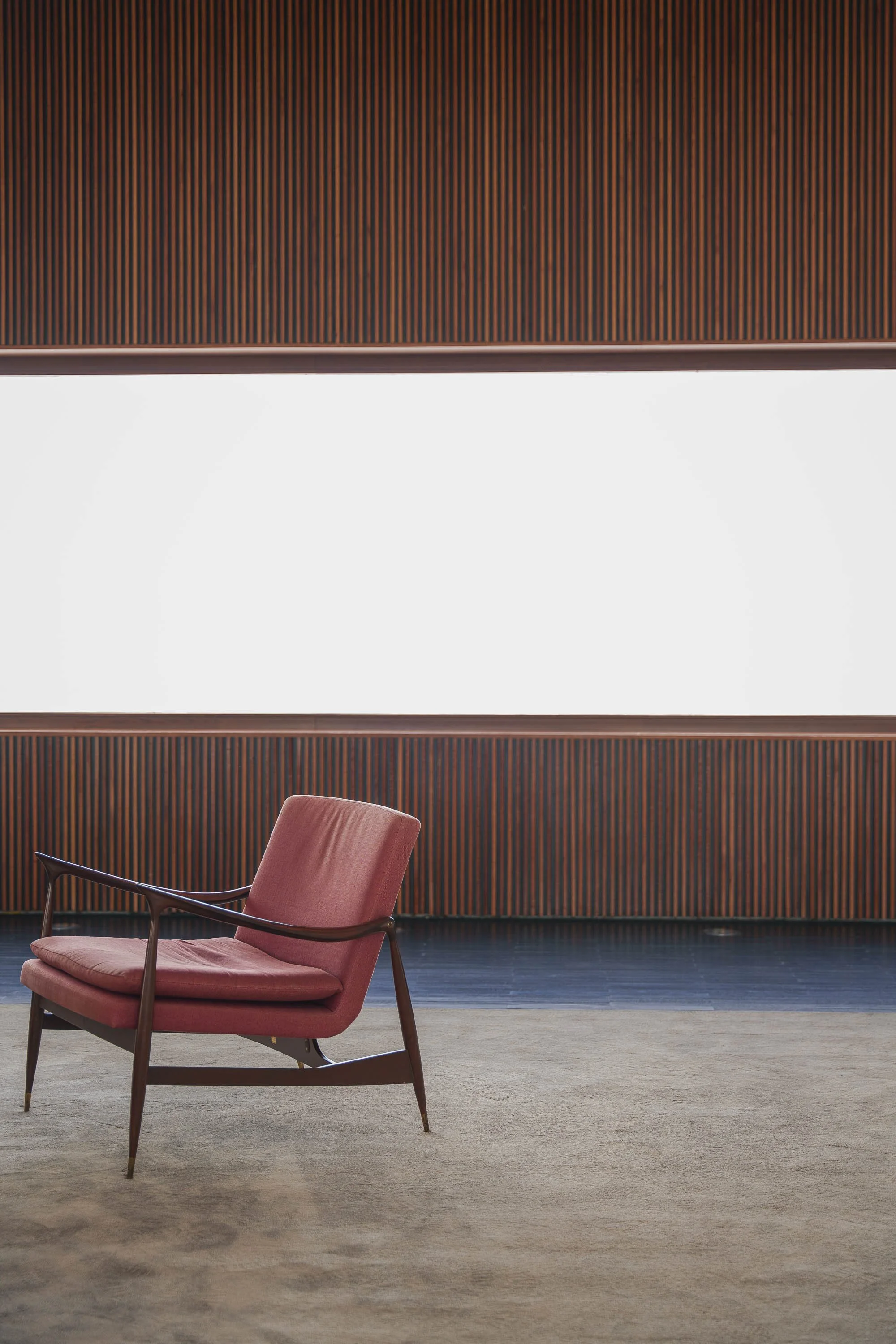 Uma sala de espera minimalista com uma cadeira de madeira com estofado rosa, parede de madeira escura vertical ao fundo, uma grande janela de vidro ao centro, e chão de carpete bege.