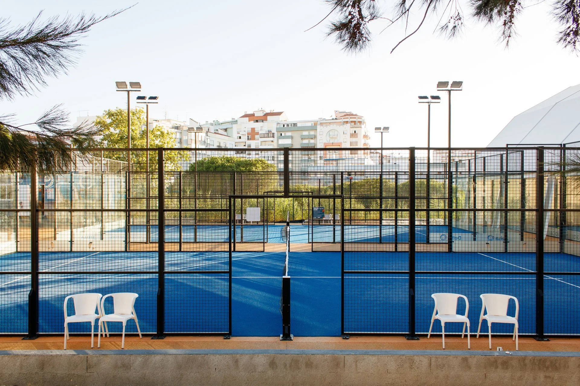 Quadra de tênis ao ar livre com redes e cadeiras brancas na frente, cercada por uma cerca de metal, com edifícios residenciais ao fundo e iluminação artificial no entardecer.