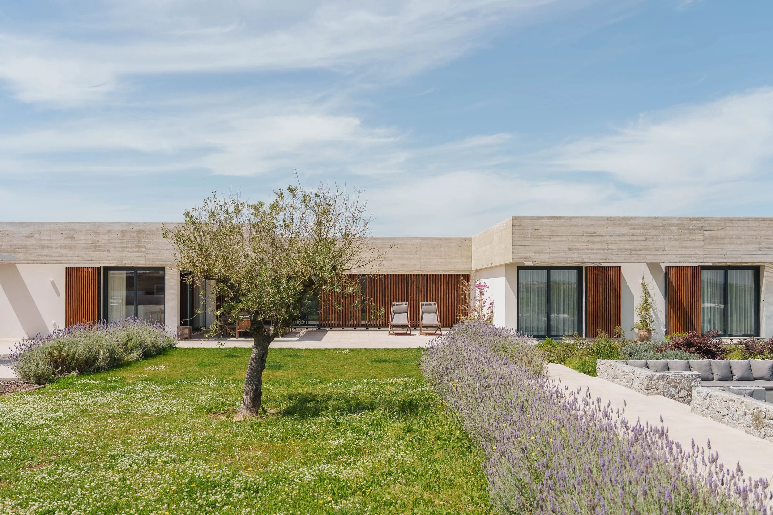 Fachada de uma casa moderna com jardim bem cuidado, árvores, arbustos de lavanda, área de descanso com cadeiras, céu azul com nuvens.