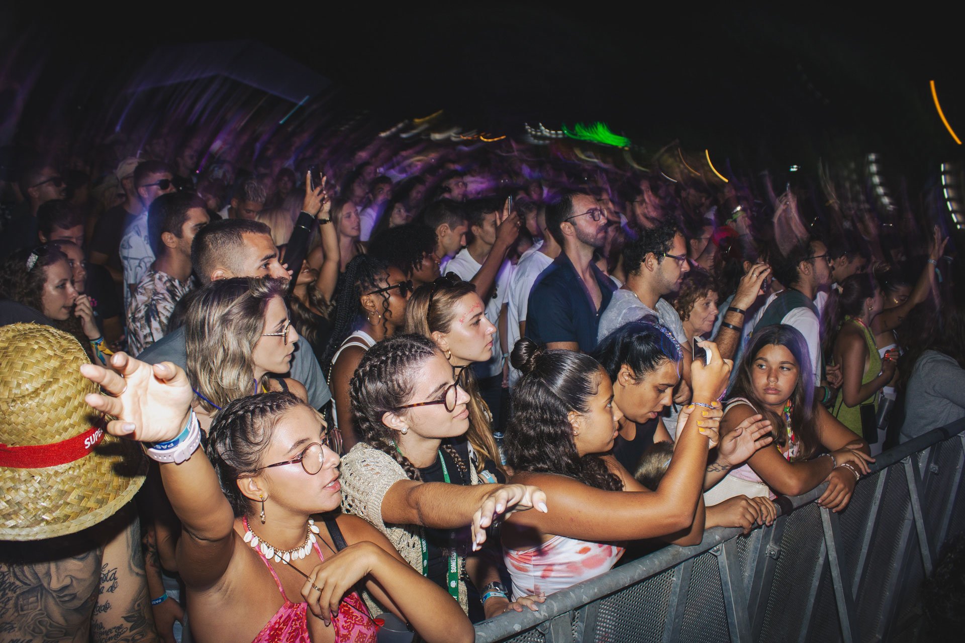 Público em um show ou evento musical, com diversas pessoas diante de uma barreira de segurança, algumas usando óculos e roupas casual. Luzes coloridas e movimento no fundo indicam um ambiente de festa ou concerto à noite.
