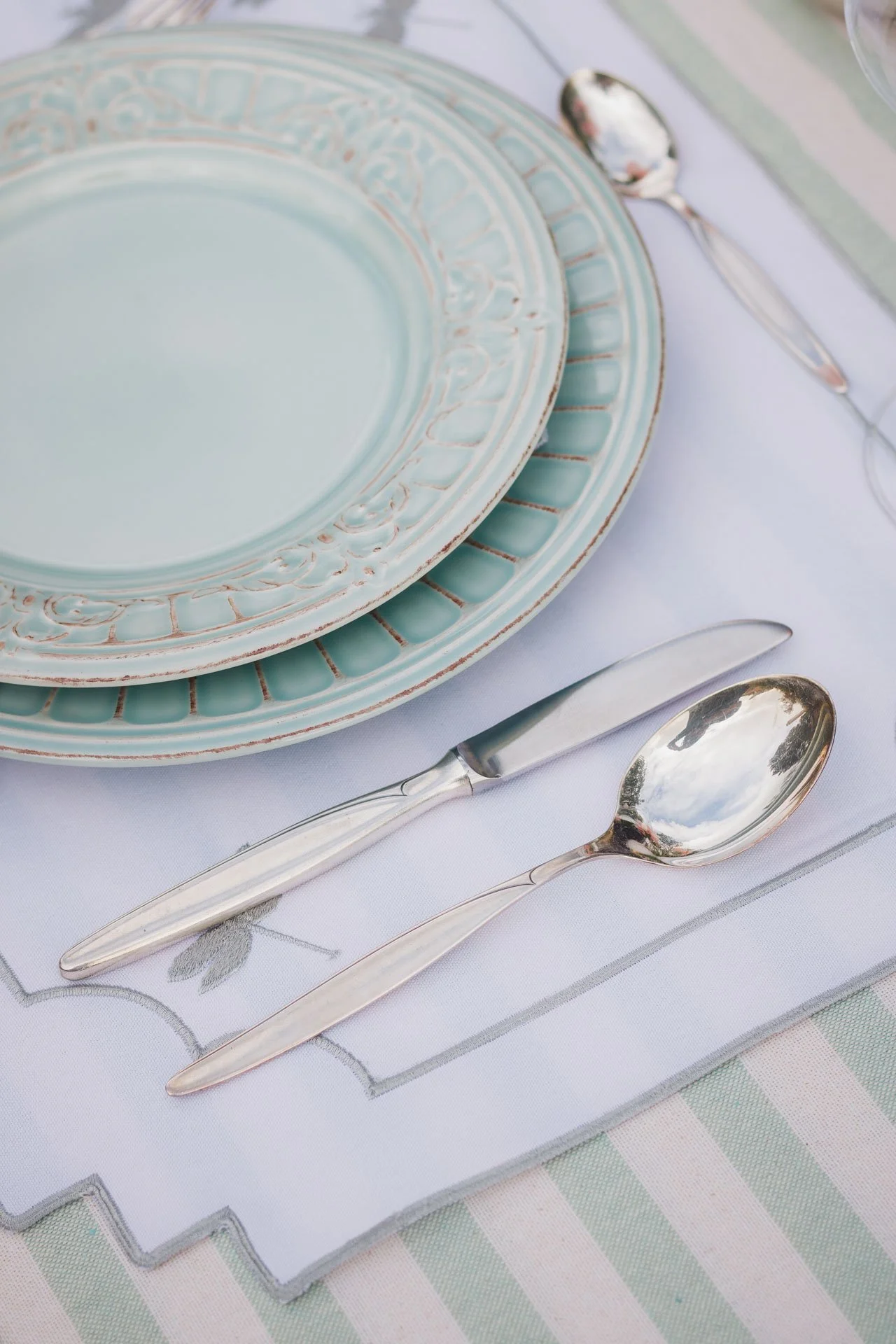 Prato de porcelana azul claro com detalhes dourados, utensílios de mesa de prata, incluindo uma faca e duas colheres, sobre uma toalha branca com bordado floral, em uma mesa com toalha de listras em tons pastel.