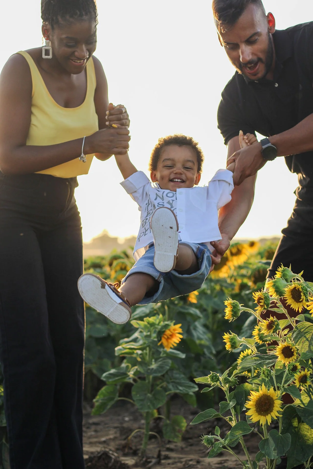 Uma criança sorridente sendo levantada por dois adultos em um campo de girassóis ao entardecer, com uma expressão de alegria.