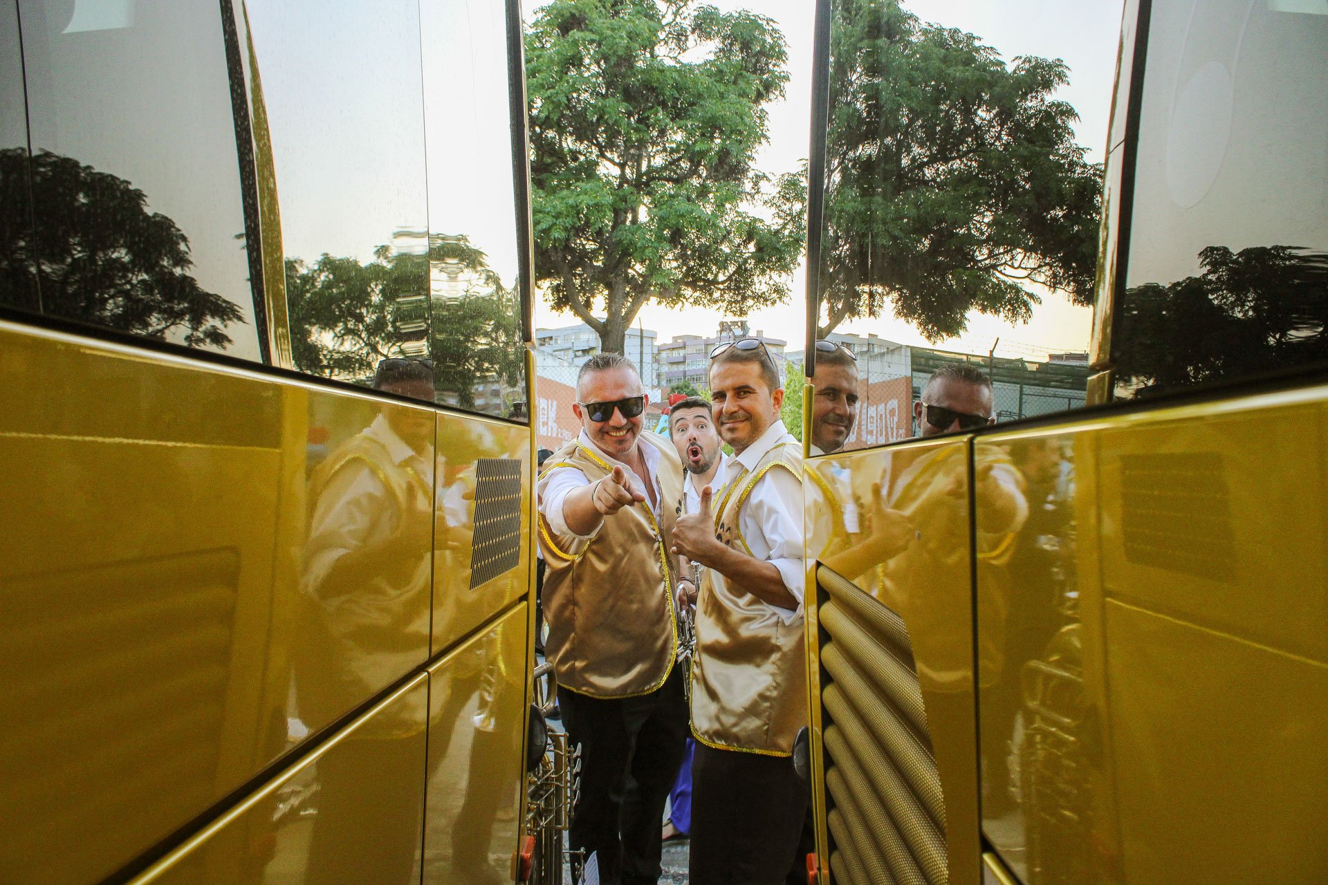 Grupo de homens sorrindo e posando, alguns usando coletes dourados e óculos escuros, ao lado de um ônibus dourado, com árvores e prédios ao fundo.