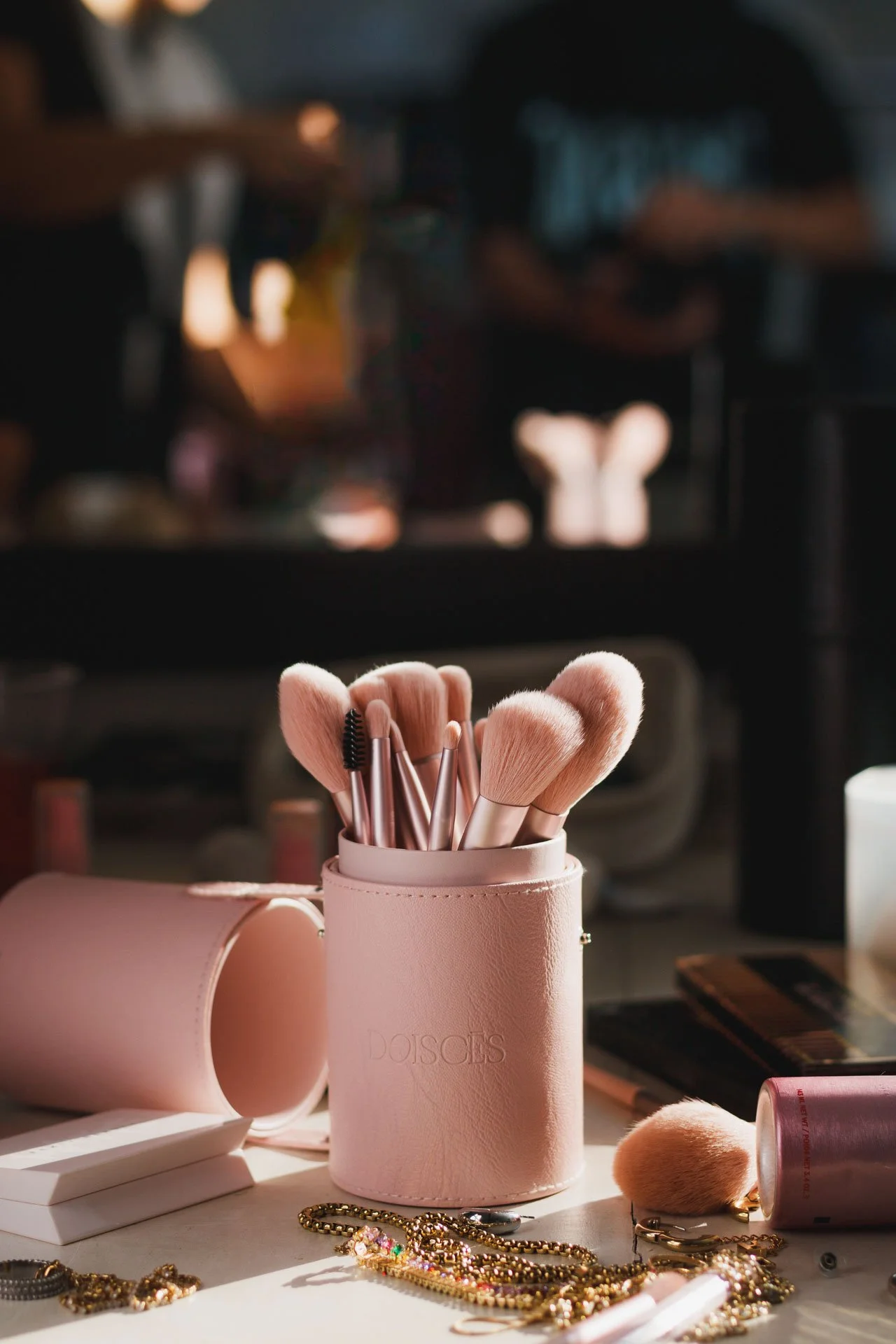 Pincéis de maquiagem em um estojo rosa claro, com acessórios como colares de pérolas e maquiagem ao redor, em uma mesa com iluminação suave.