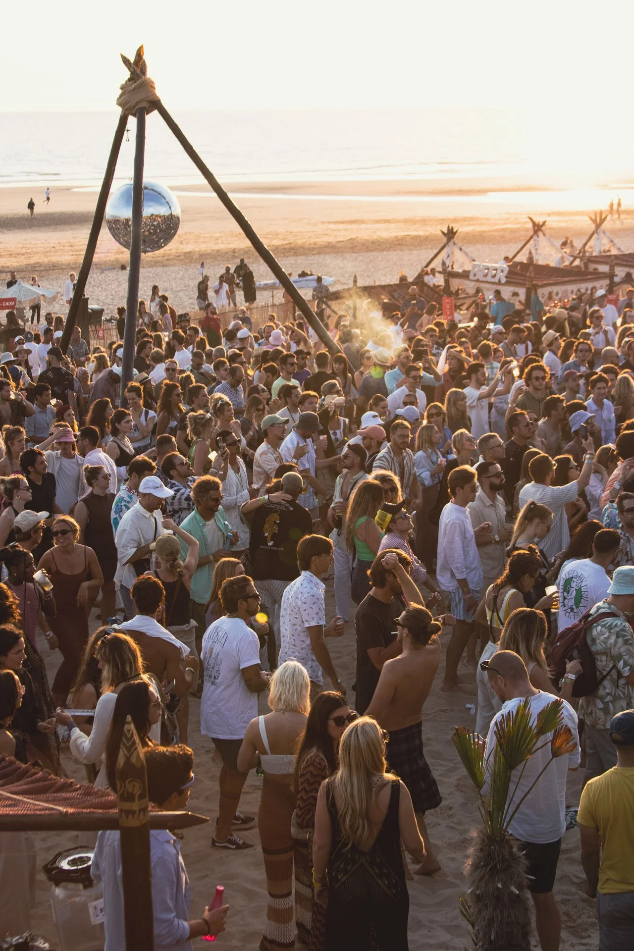 Multidão de pessoas na praia ao pôr do sol, com várias barracas e uma escultura de espelho no fundo.