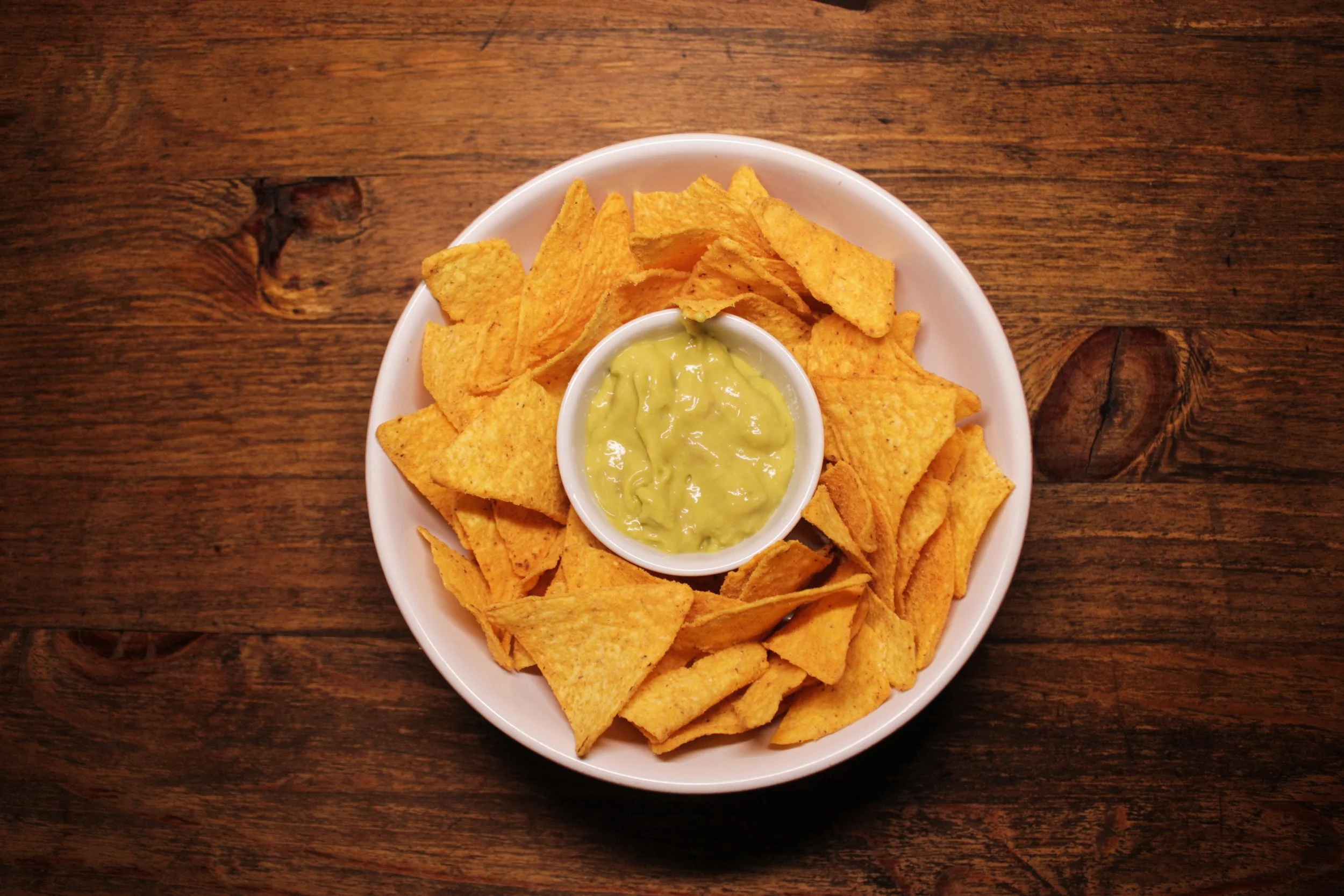Prato com nachos de milho cobertos por molho de queijo na tigela central, sobre uma tábua de madeira.