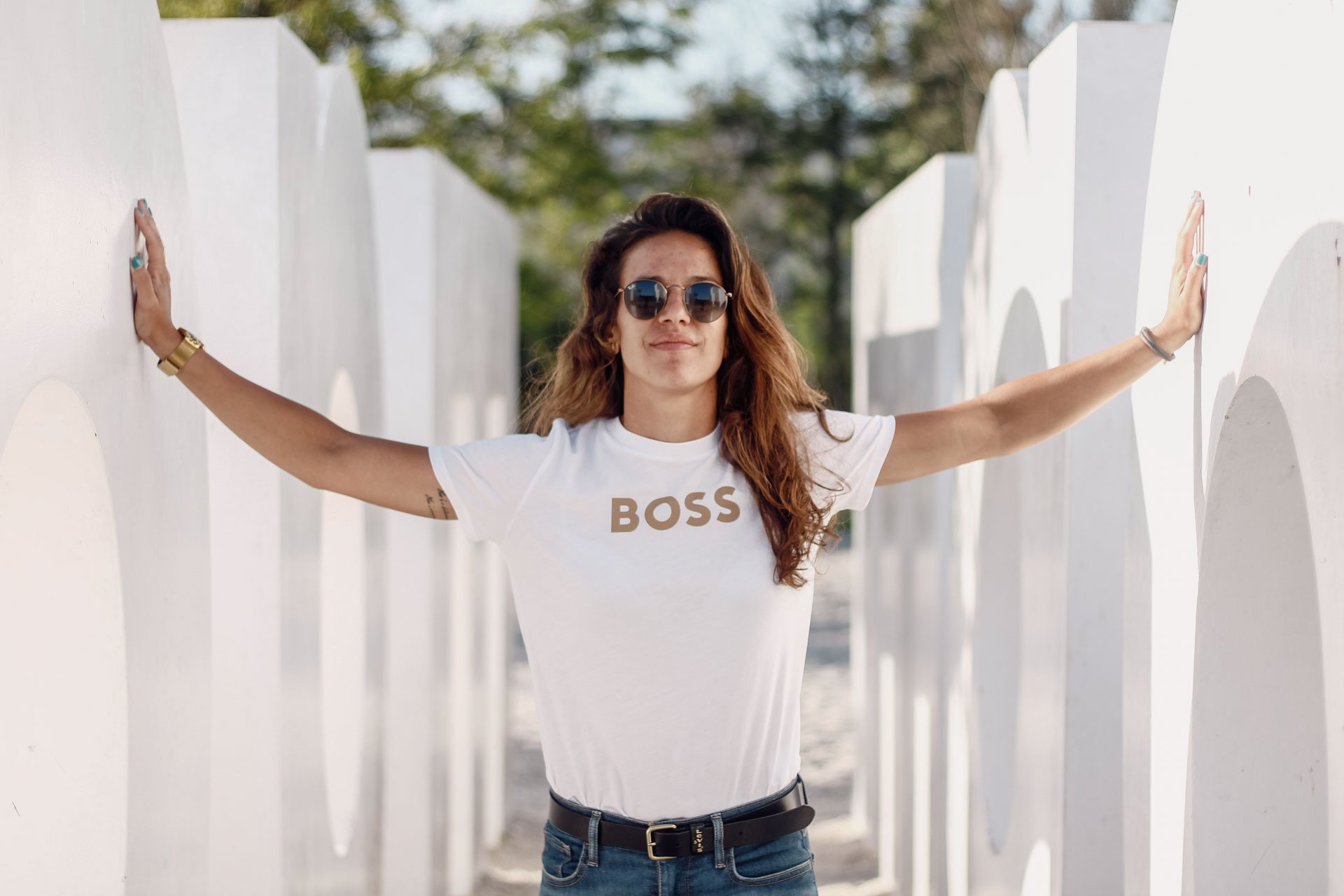 Jovem mulher com cabelos cacheados, óculos escuros, camiseta branca com a palavra 'BOSS', jeans e pulseira, posicionada entre duas paredes brancas com desenhos, arms abertos tocando ambas as paredes.