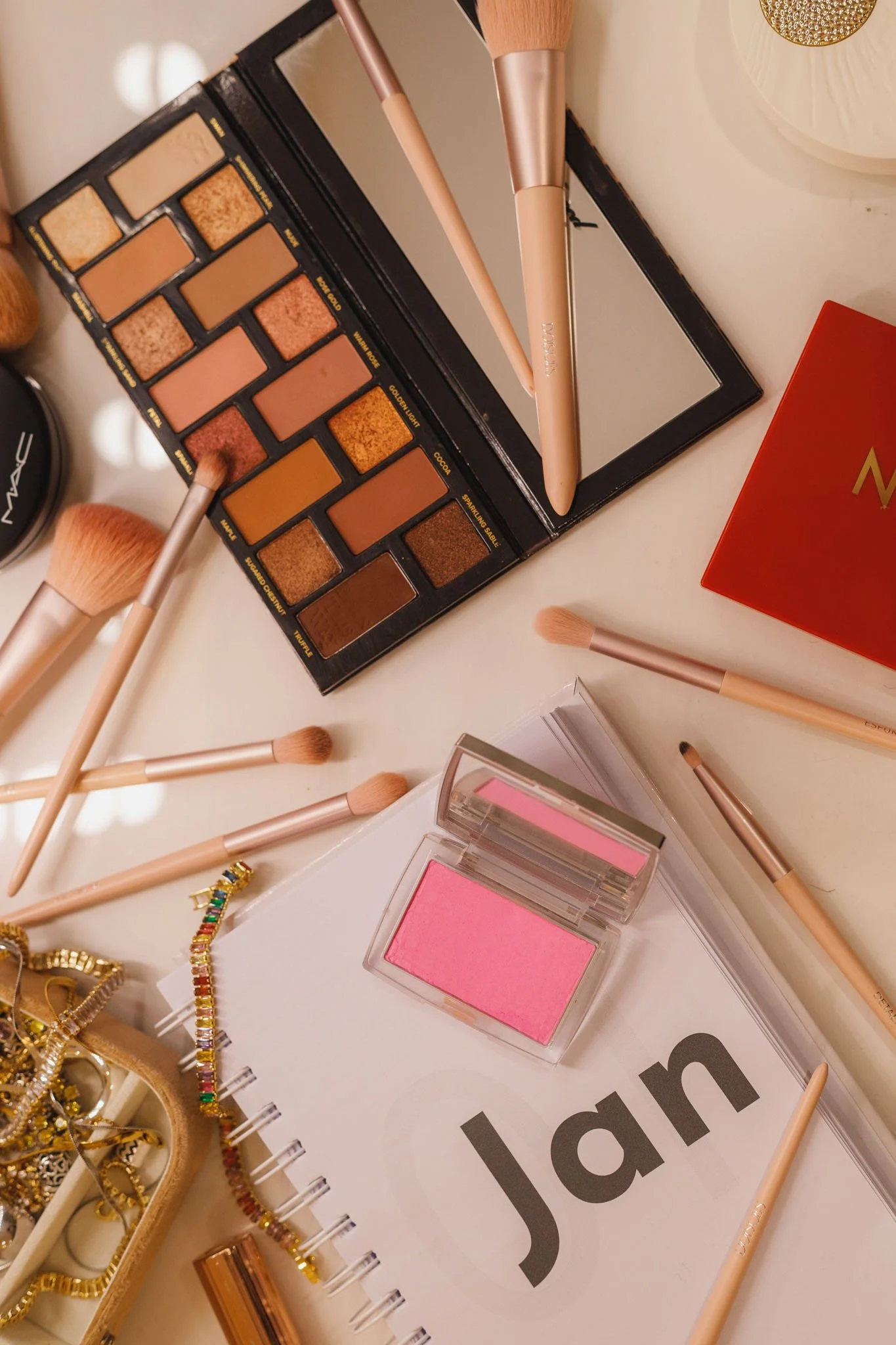 Maquiagem com paleta de sombras, pincéis, blush rosa, acessórios dourados, um caderno com a data 'Jan' e uma caixa com joias.