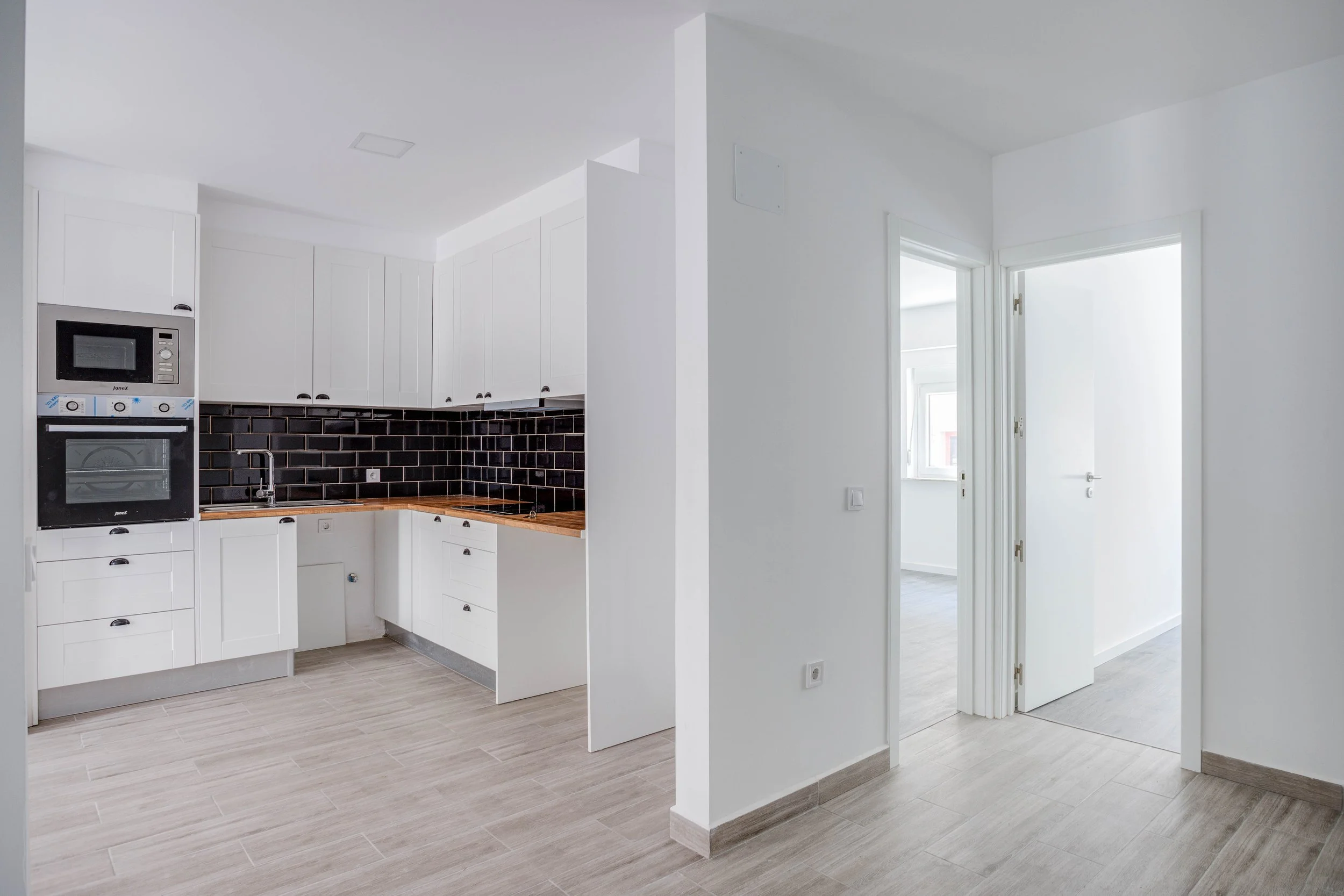 Cozinha moderna com armários brancos, bancada de madeira, backsplash de azulejos pretos e eletrodomésticos embutidos, vista para uma área com janelas e portas brancas