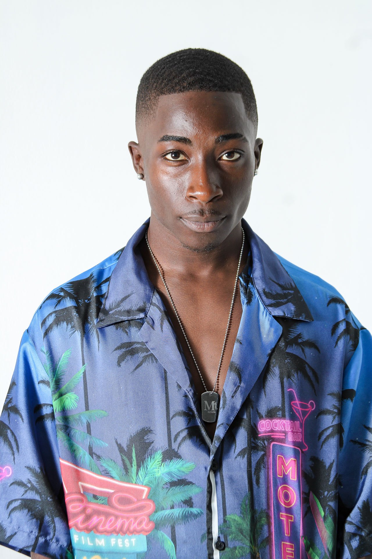 Homem com cabelo curto e escuro, usando uma camisa azul com estampa de Palmeiras e neon com temas de bar de coquetel, com colar prateado e brincos pequenos, olhando diretamente para a câmera.