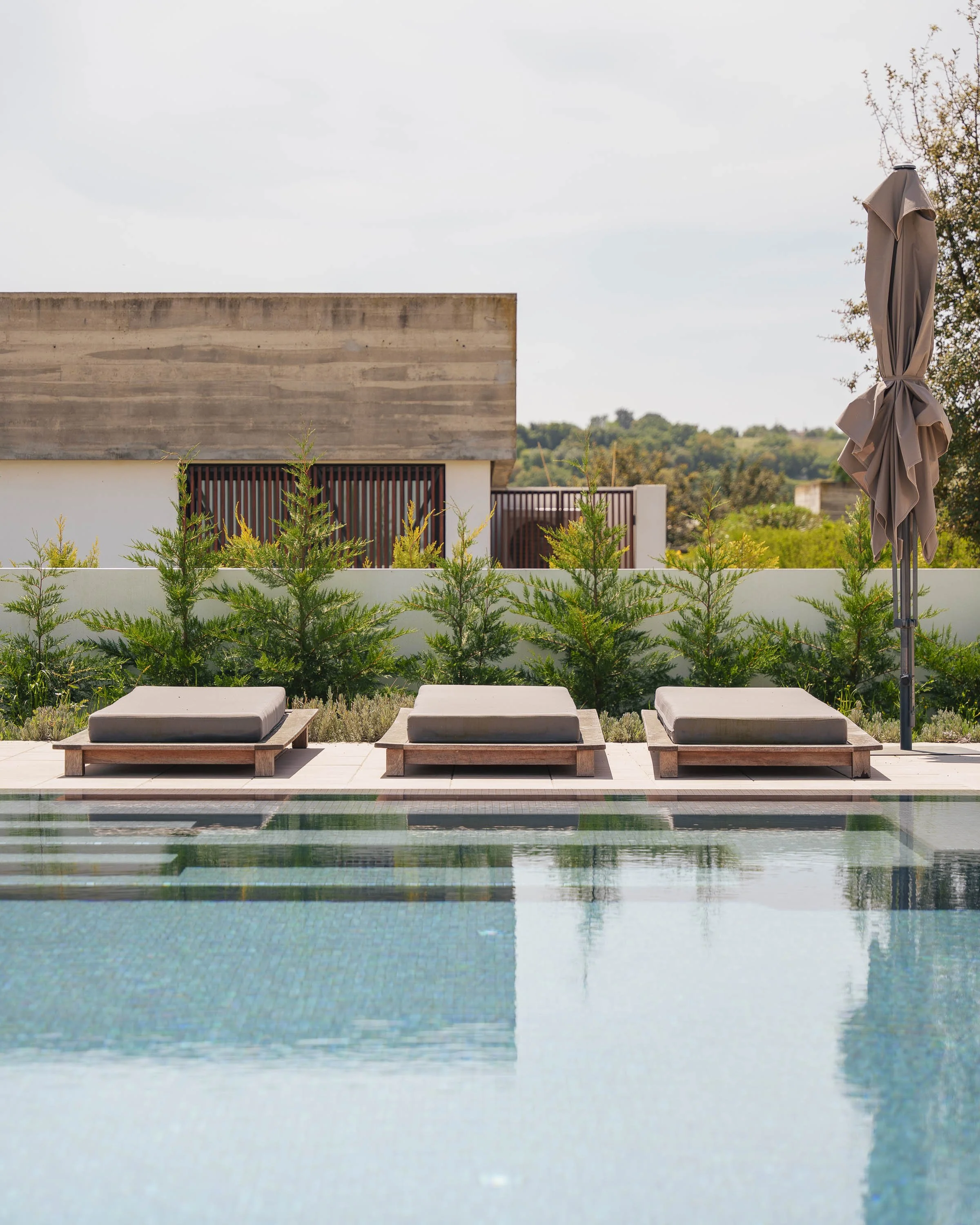 Três cadeiras de piscina com almofadas ao lado de uma piscina, cercadas por arbustos verdes e uma parede branca, com um prédio moderno ao fundo e um guarda-sol fechado à direita.