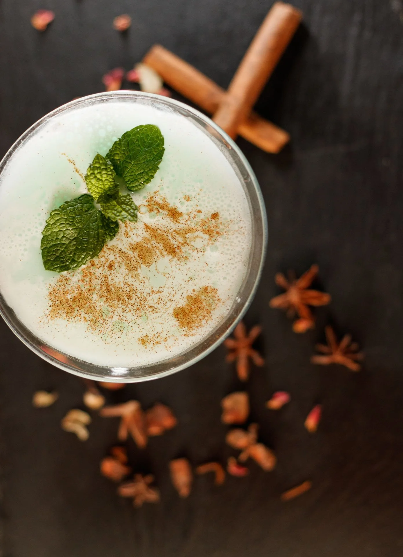 Copo de bebida com folhas de hortelã e canela em pó, sobre uma mesa escura com pedaços de canela em pau, sementes de cardamomo e estrelas de anis.