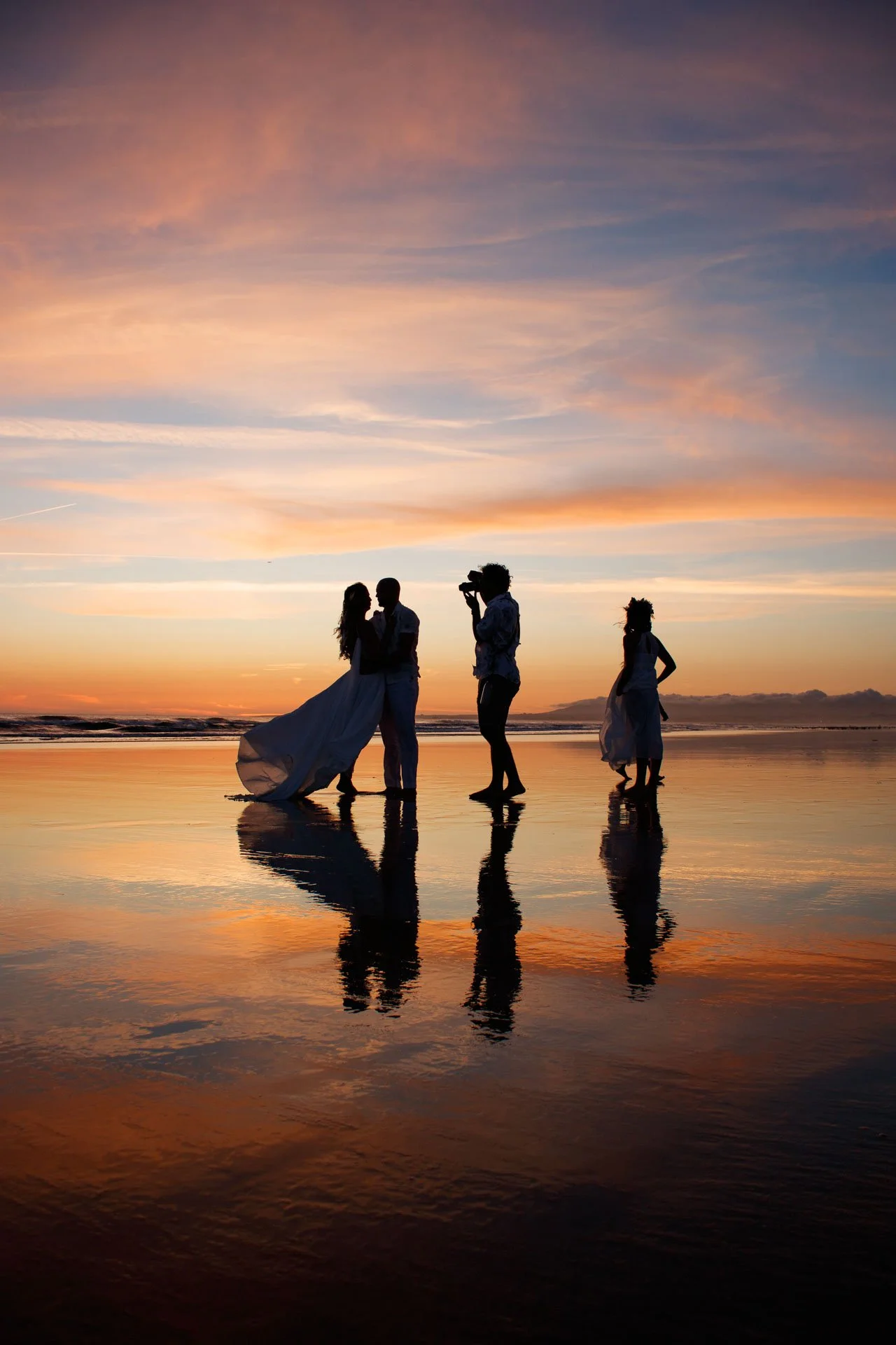 Silhuetas de uma celebração na praia ao pôr do sol, com um casal de noivos e duas pessoas ao fundo, uma delas tirando fotos.