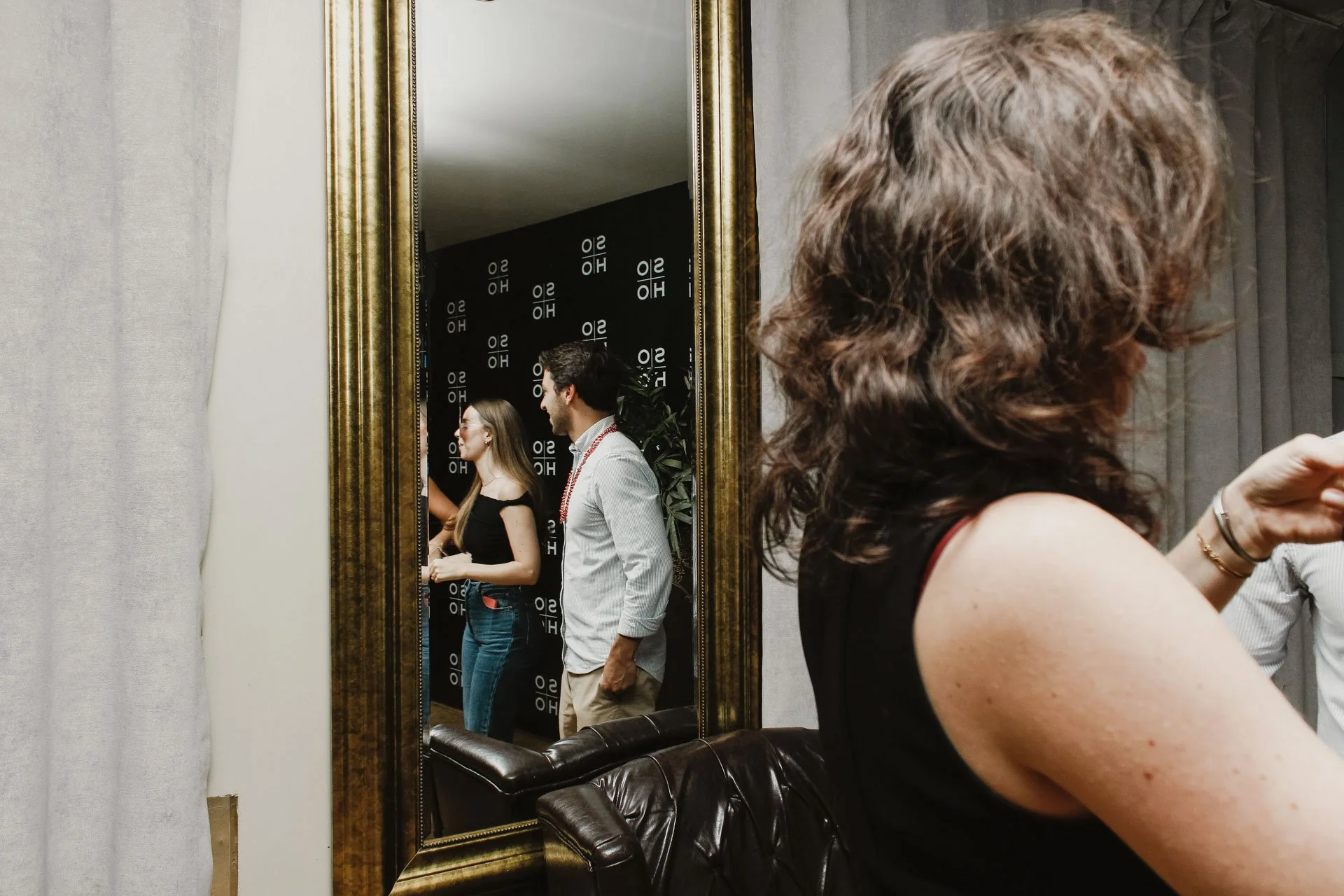 Pessoas refletidas em um espelho grande, com fundo preto com logos e textos em branco, mostrando duas mulheres e um homem em uma sala de evento.