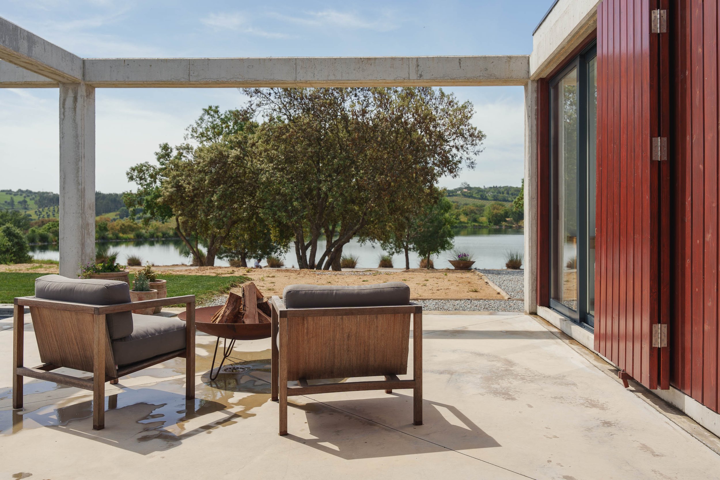 Sacada de casa com duas poltronas de madeira e almofadas cinzas, vista de árvores e rio ao fundo.