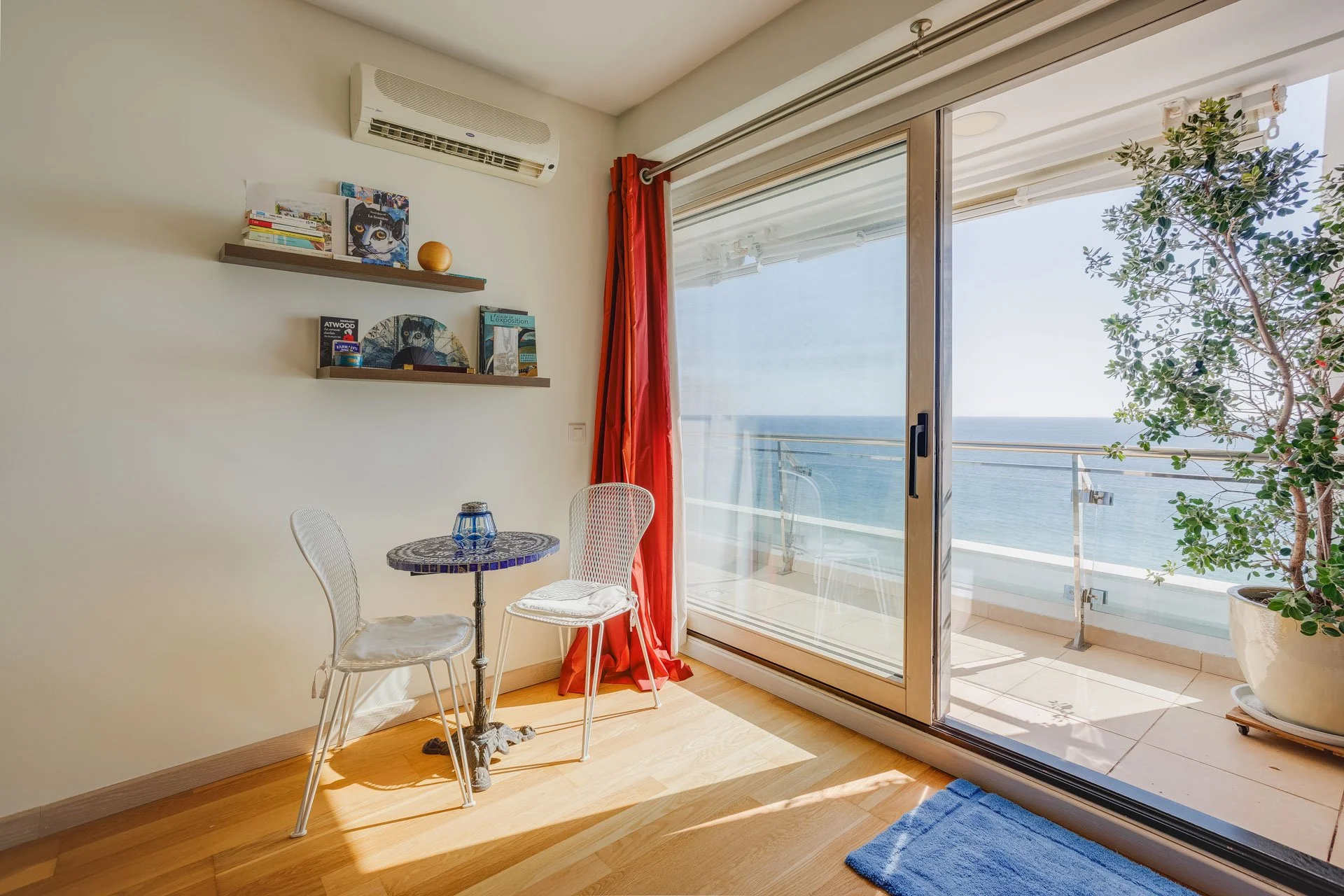 Sala de estar com mesa de bilhar, cadeiras brancas, cortina vermelha, varanda com vista para o mar, e planta em vaso grande.