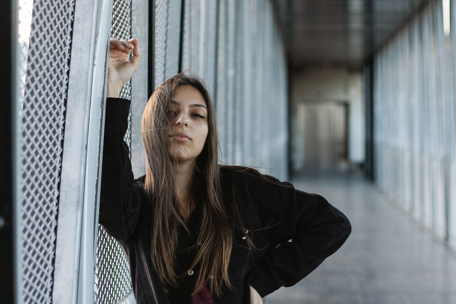 Jovem mulher posando na passarela de um ambiente industrial, com paredes de metal e na diagonal, usando casaco preto.